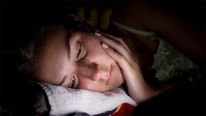 a gif of a woman in a darkened room in bed with her face lit up by a mobile phone
