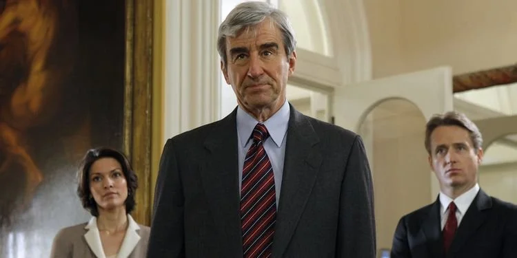 Sam Waterston in suit and tie in courthouse with Alana De La Garza and Linus Roache standing in background.