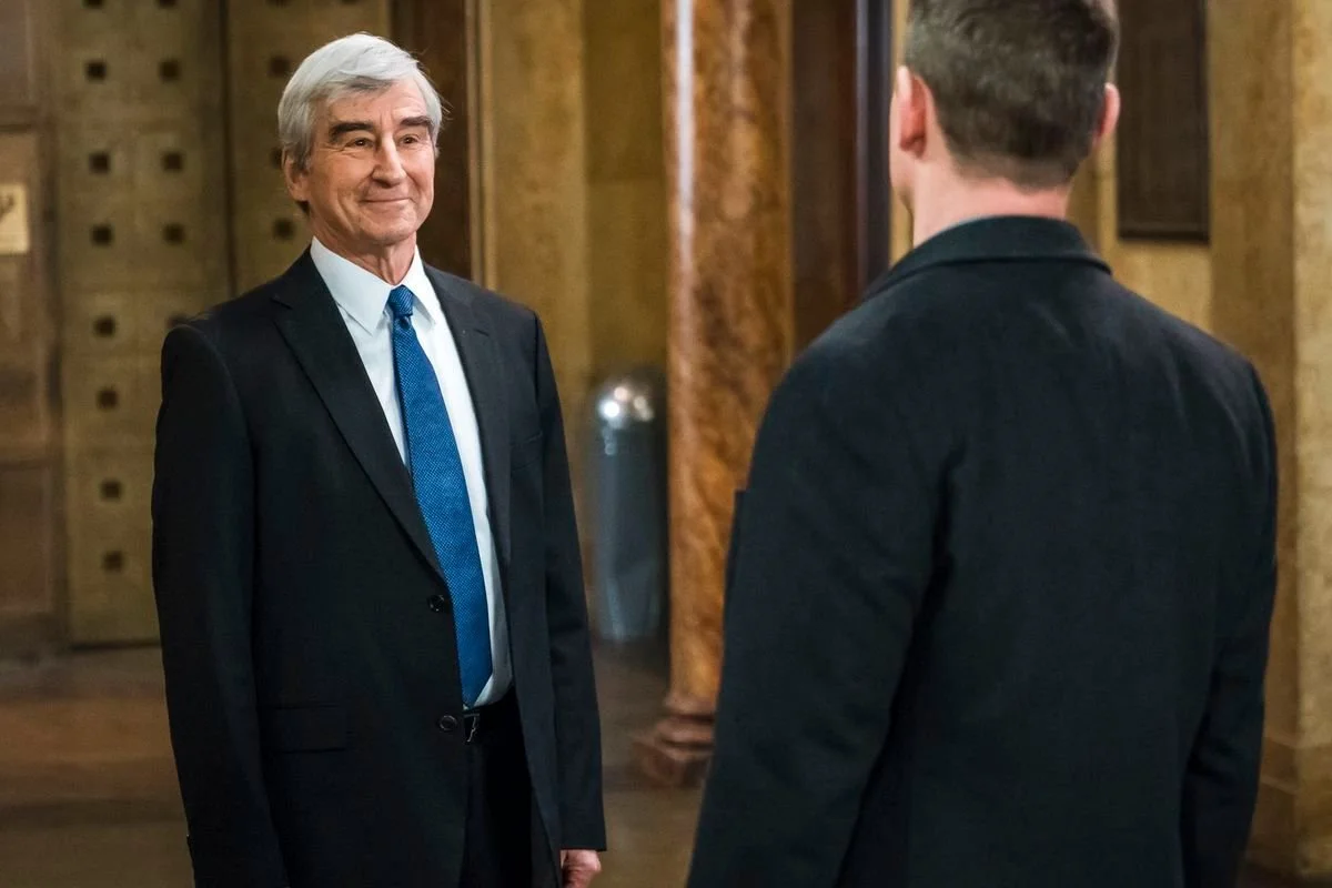 Sam Waterston as Jack McCoy in dark suit and tie standing in a marble lobby with a man's back in the foreground