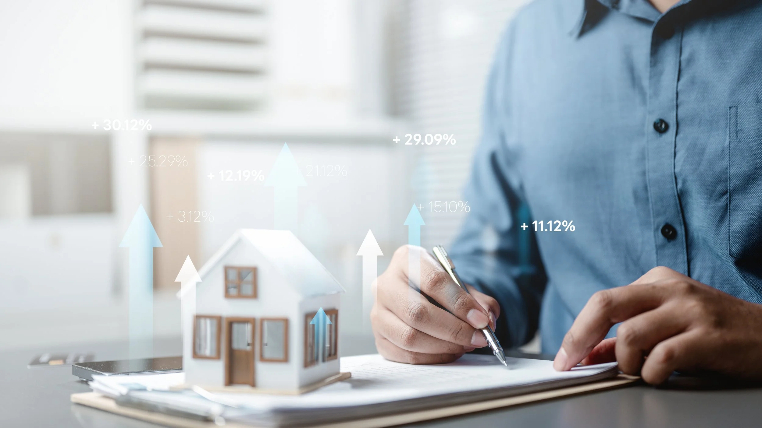 A person in a blue shirt holding a pen and writing on paper, with digital financial data and graphs overlaid, and a small model house on the table.