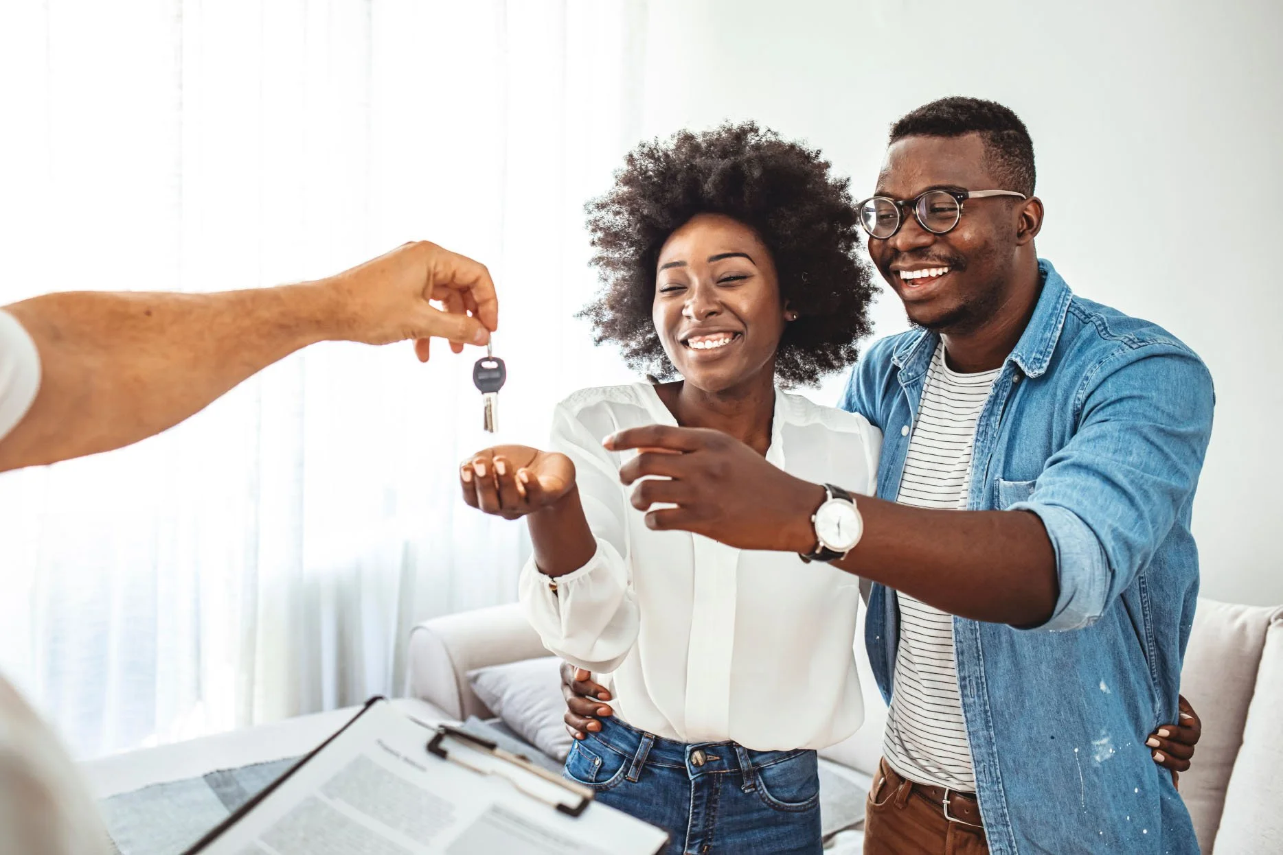 A happy couple is exchanging keys during a home purchase or rental process, indoors with natural light. The woman has curly hair and is smiling while holding the keys, and the man, wearing glasses and a denim shirt, is smiling and assisting her.