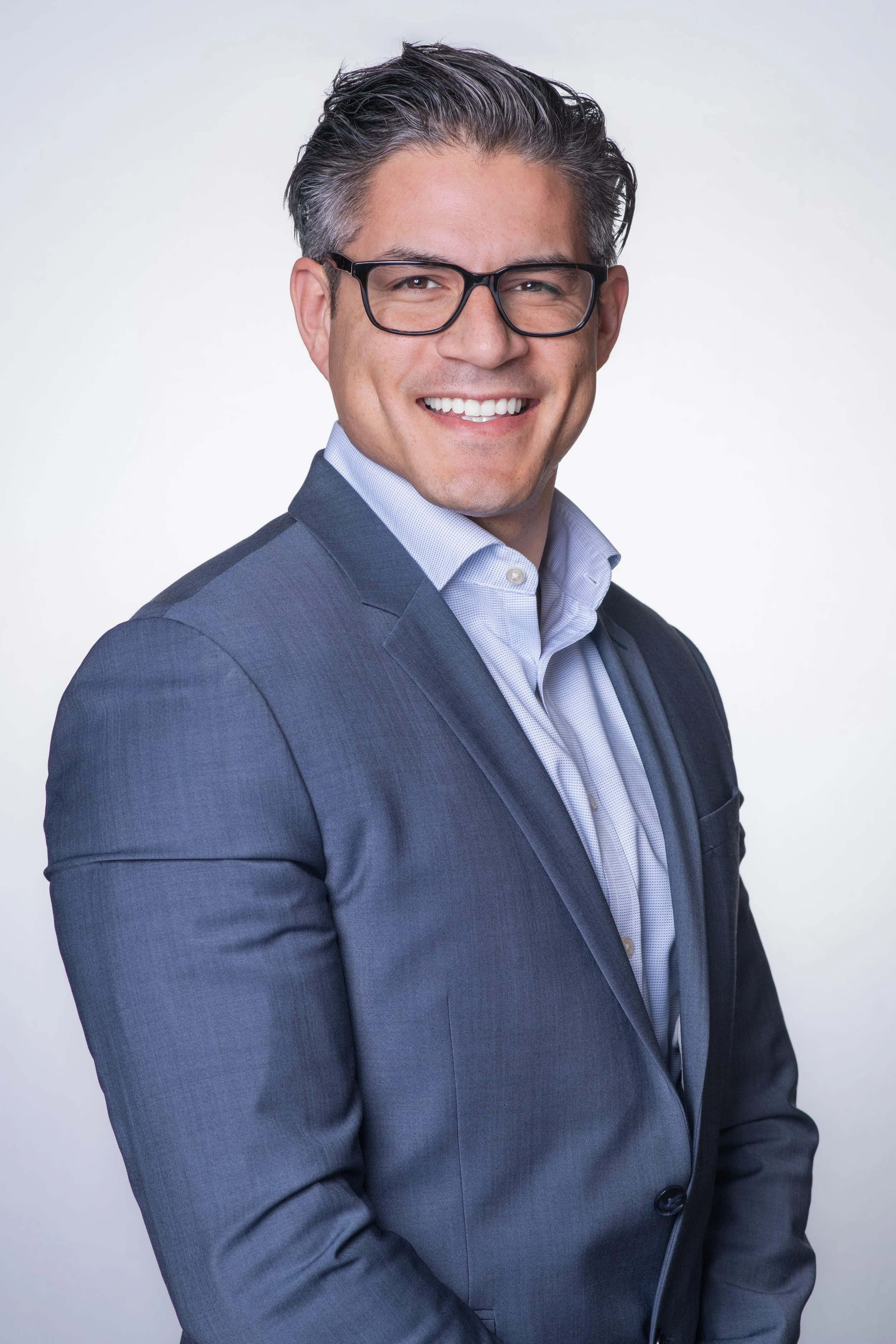 Professional headshot of a man wearing a blue suit and glasses with a white backdrop. He is facing camera right.