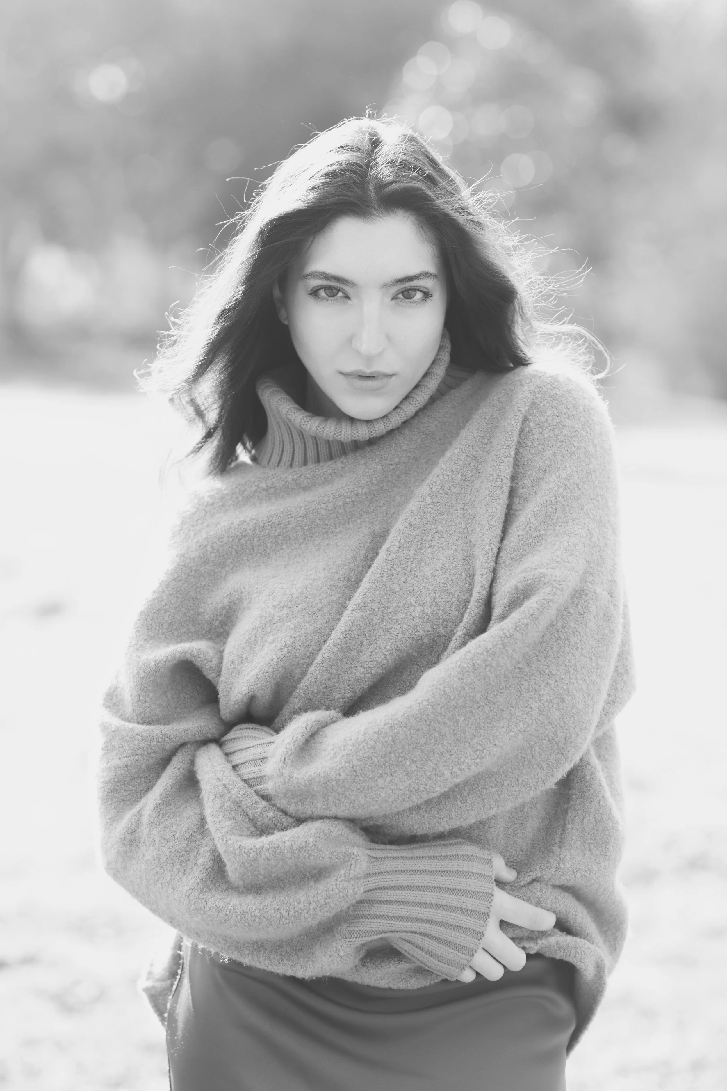 Black and white portrait of a young woman outdoors, wearing a cozy sweater with a high collar and long sleeves, looking directly at the camera with her arms crossed.