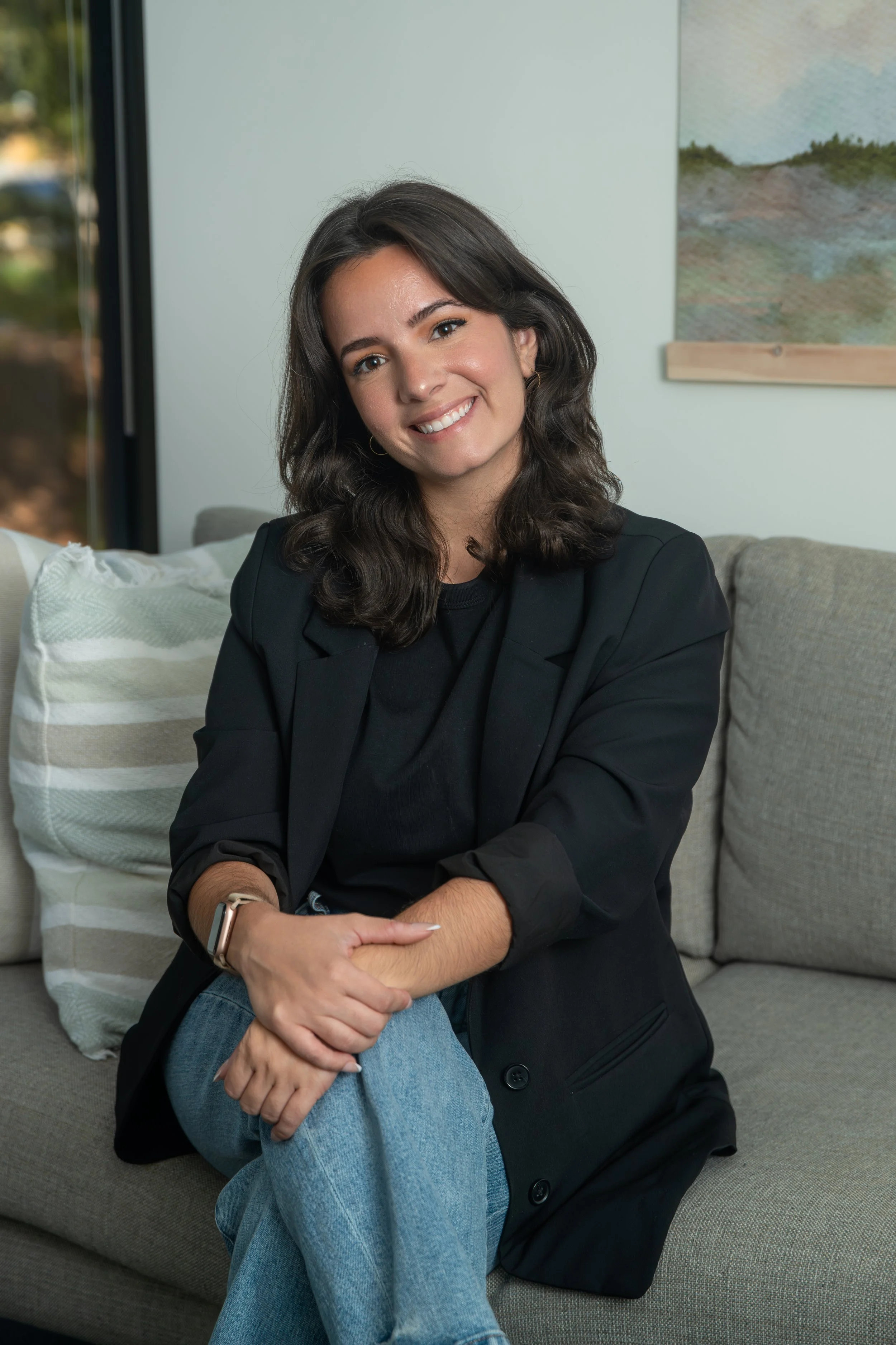 A young woman with shoulder-length dark hair, smiling, sitting on a light-colored sofa with striped pillows, wearing a black blazer, black shirt, blue jeans, and a watch, in a room with a window and a wall painting.