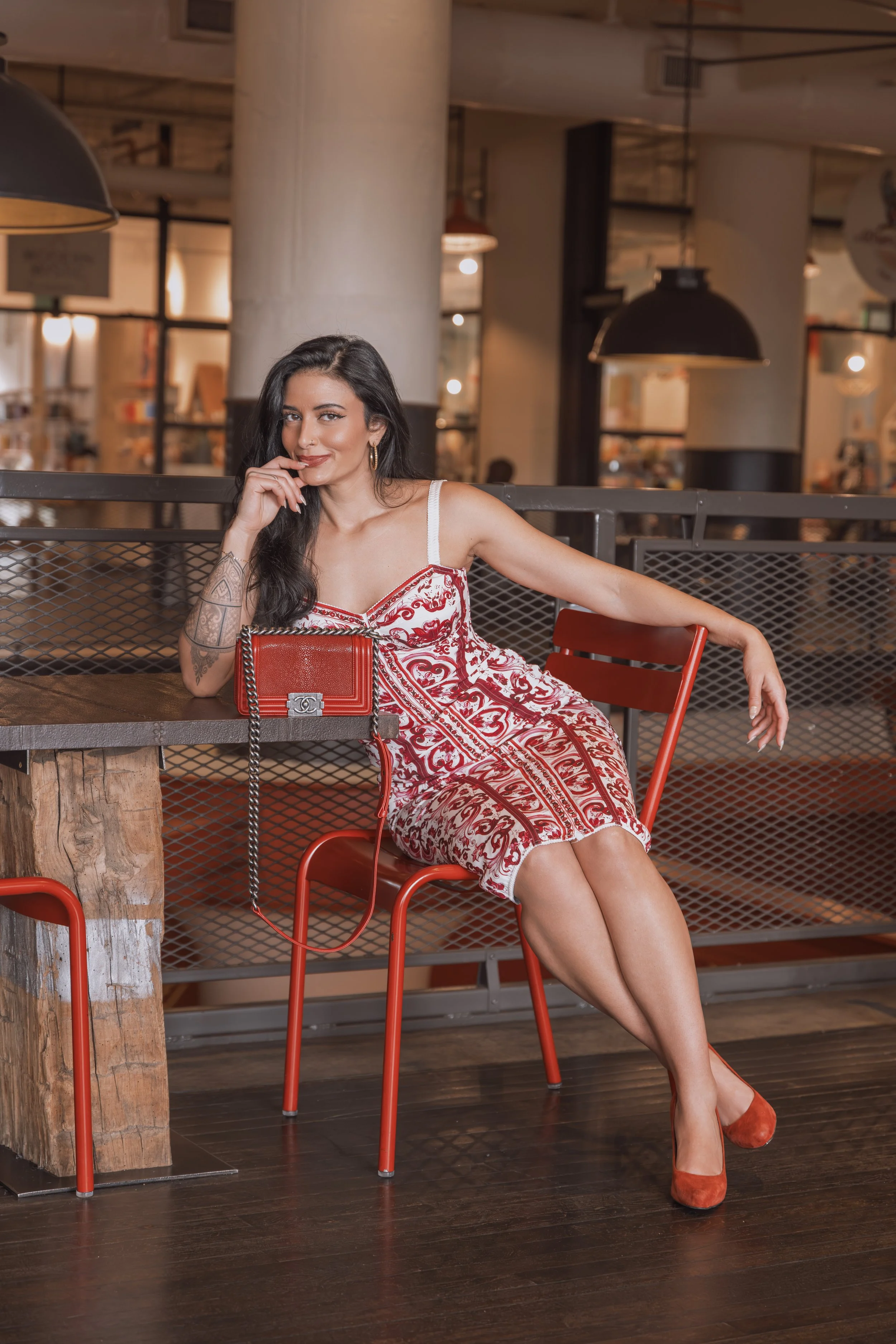 A woman with long dark hair sitting on an orange chair at a wooden table in a rustic outdoor cafe with warm lighting, wearing a red and white patterned dress and red shoes, with a red purse on the table.