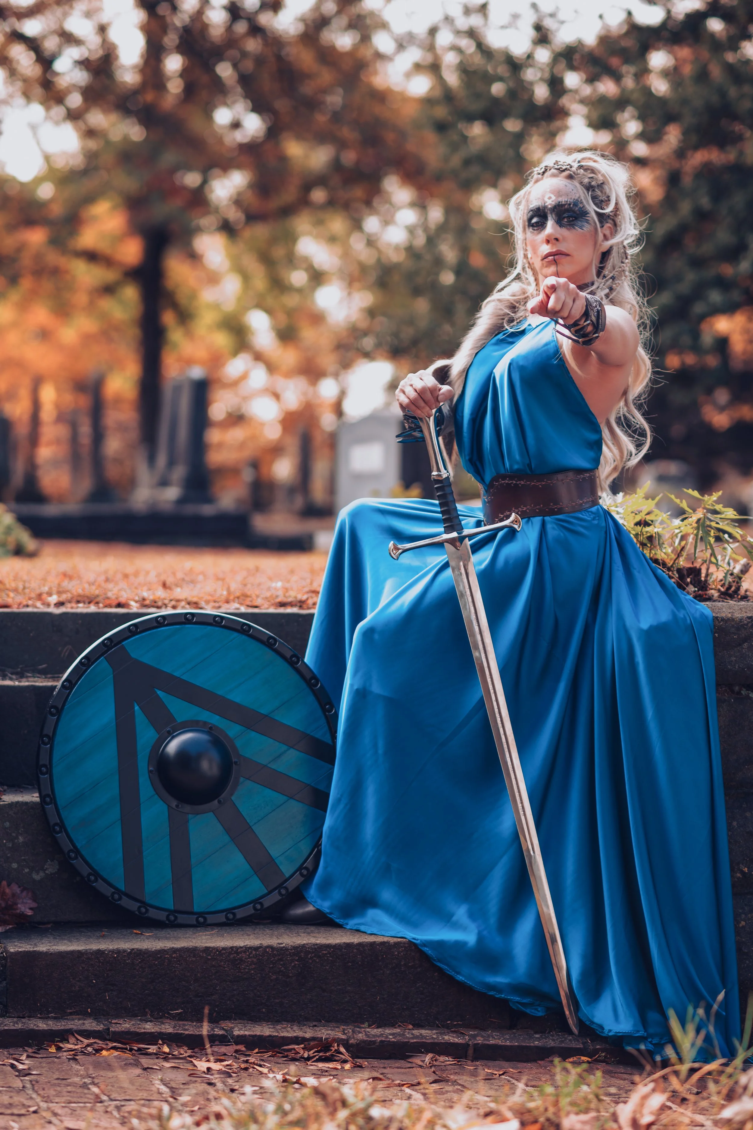 Woman dressed as a warrior princess in a blue dress holding a sword and shield, sitting outdoors with autumn foliage in the background.