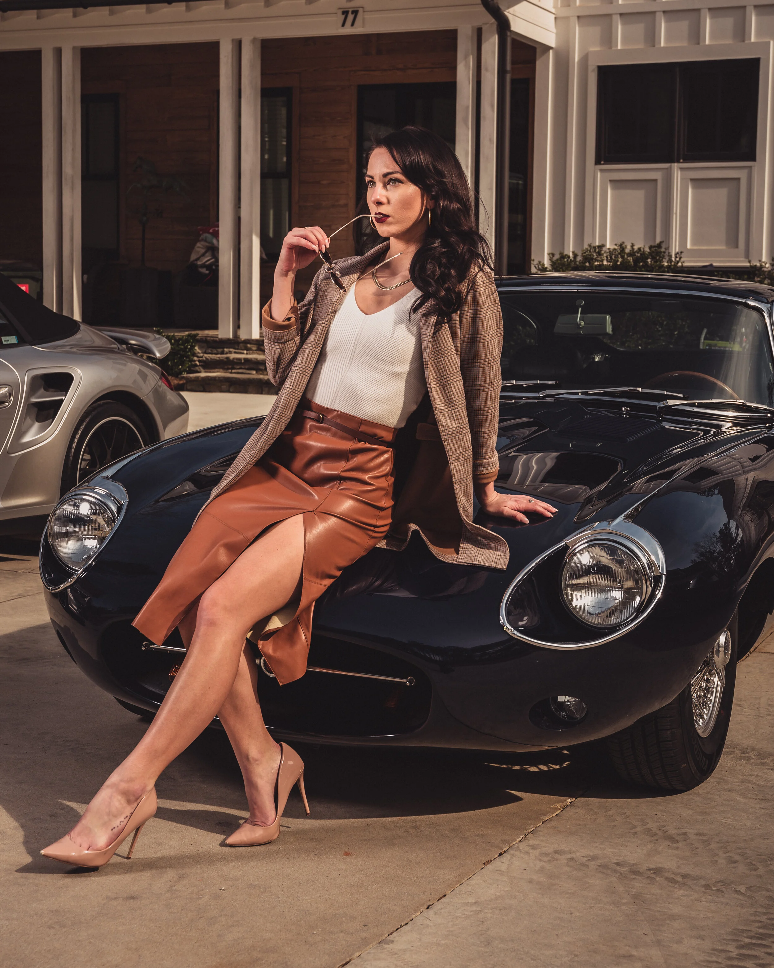 A woman dressed in a white top, leather skirt, and blazer sitting on the hood of a black sports car.