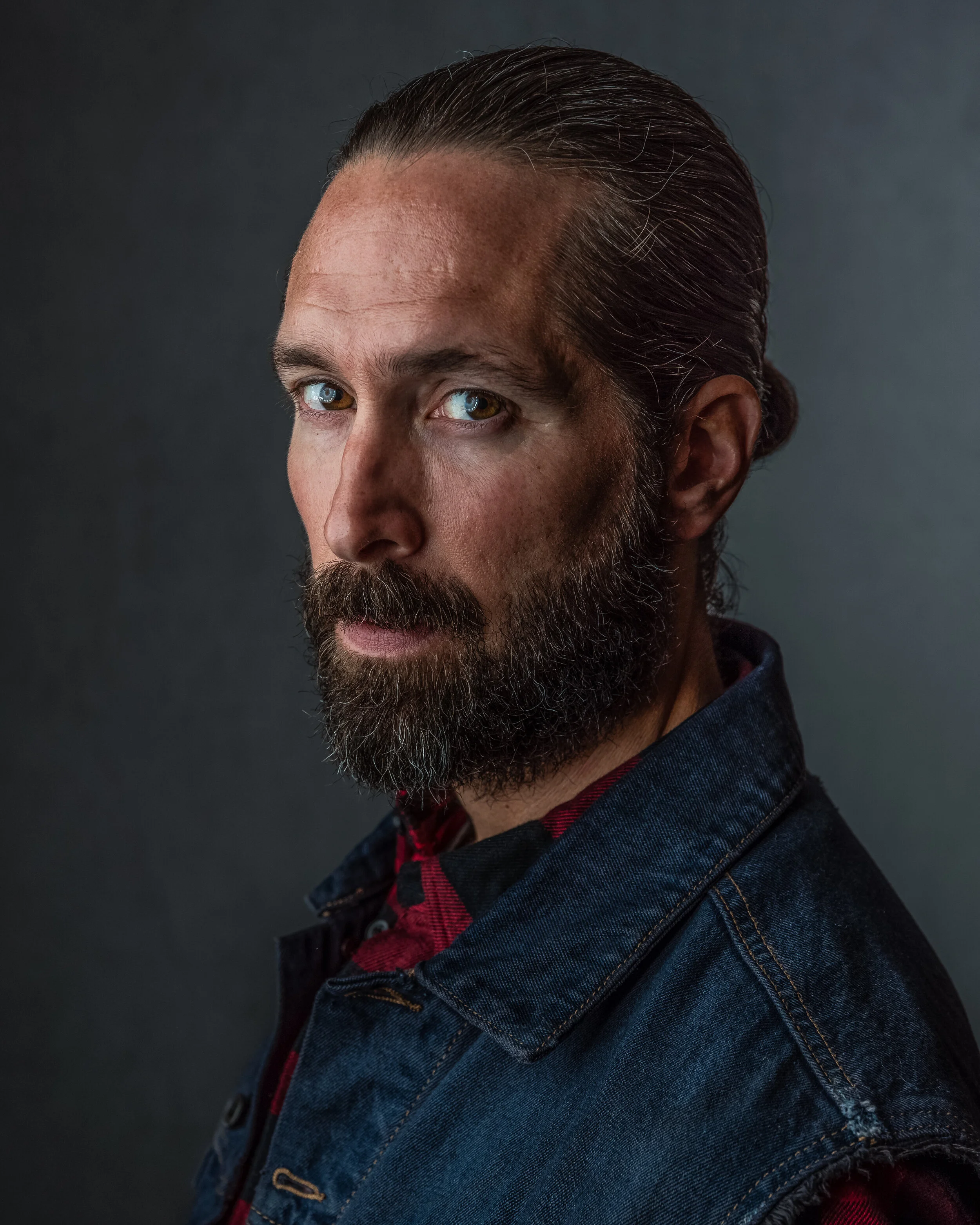 A man with long, slicked-back hair and a thick beard, wearing a denim jacket over a red and black checkered shirt, looking over his shoulder against a dark background.
