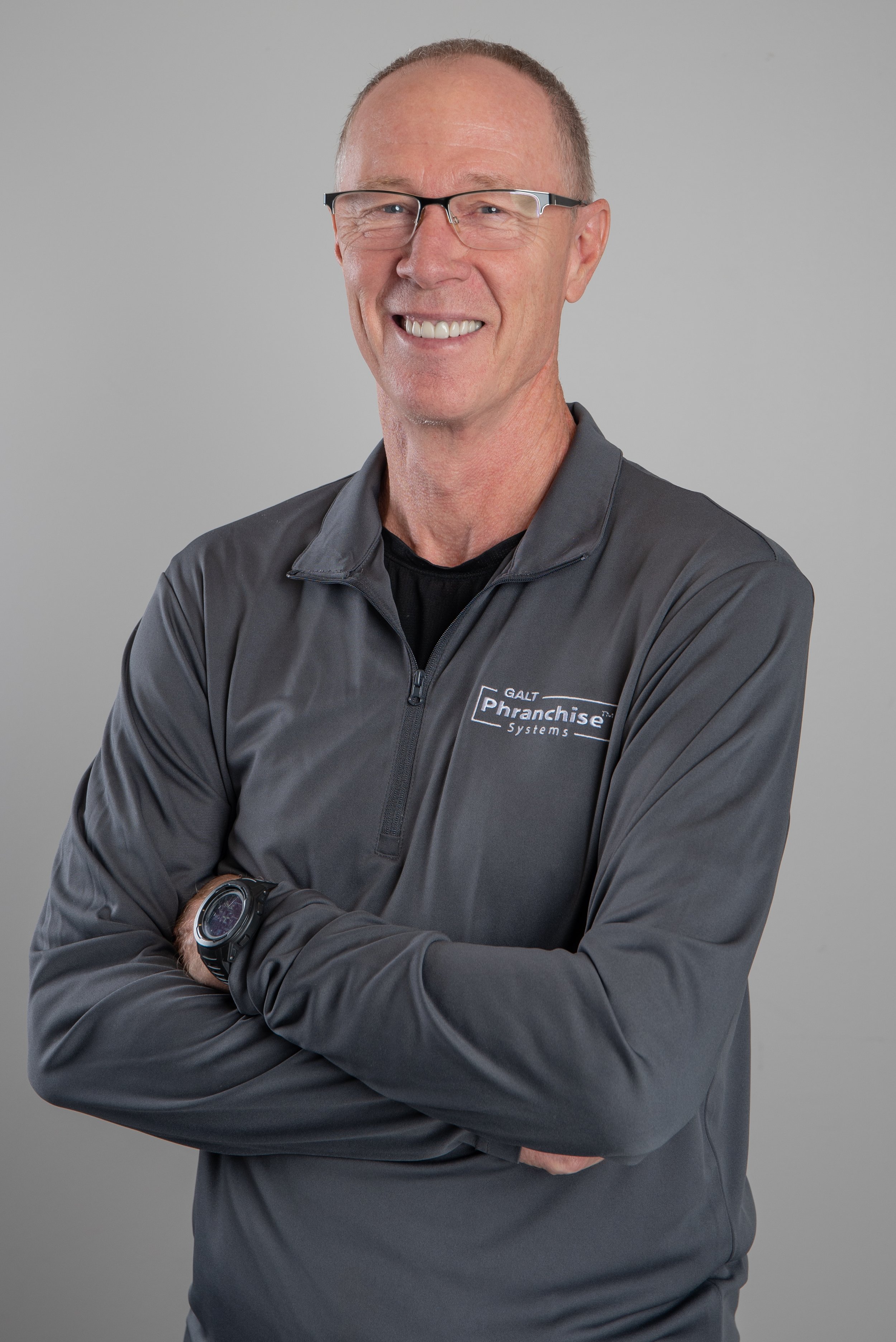 A smiling man with short hair and glasses wearing a gray quarter-zip jacket with a logo, standing with arms crossed against a plain gray background.