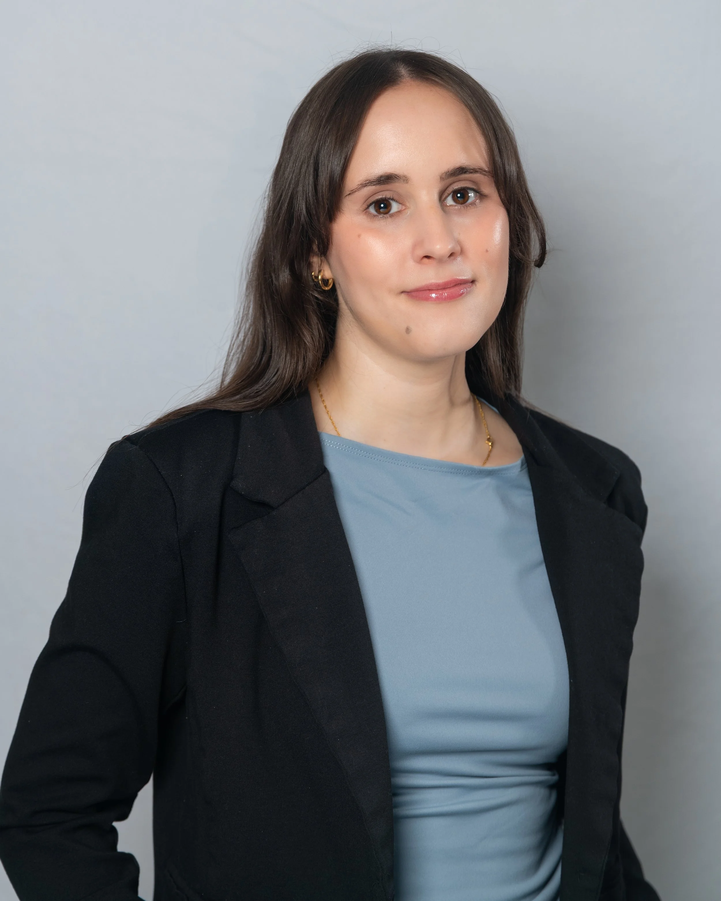 A woman with shoulder-length brown hair wearing a black blazer, light blue top, and gold jewelry, standing against a plain light gray background.