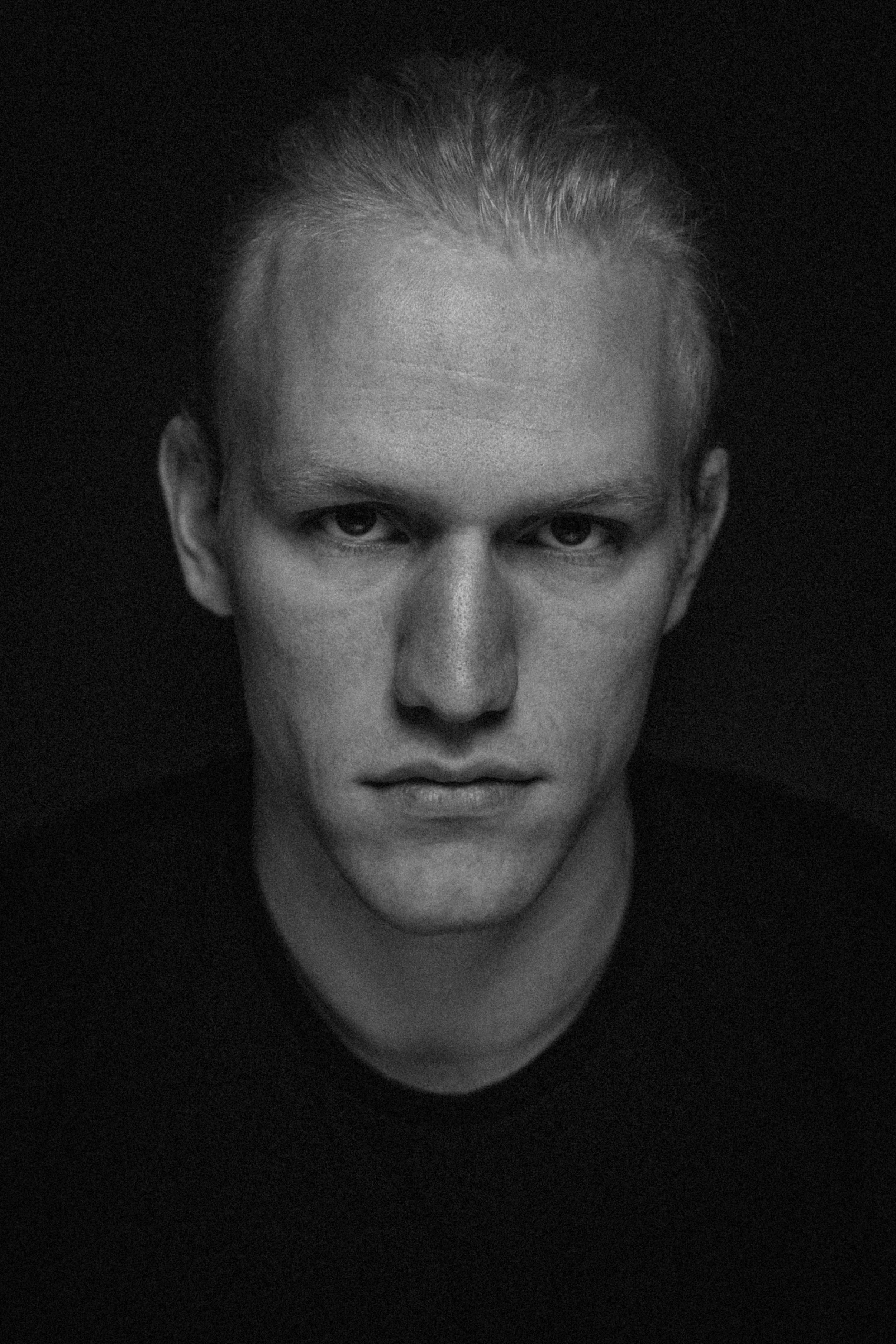 A black and white portrait of a young man with light skin, light hair pulled back, staring directly at the camera with a serious expression.
