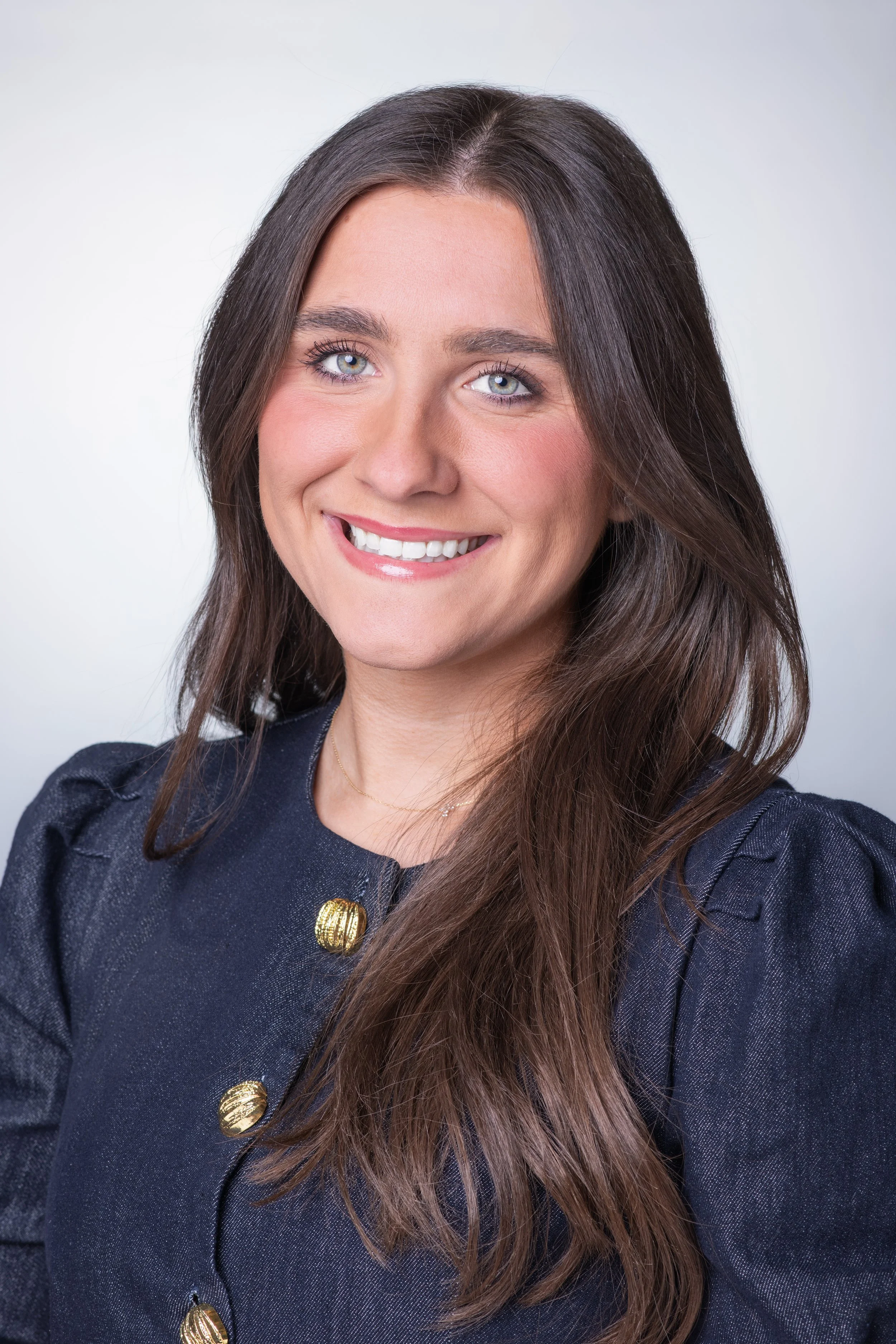 Professional headshot of a woman with a white backdrop wearing a blue top and smiling. She is facing camera right.