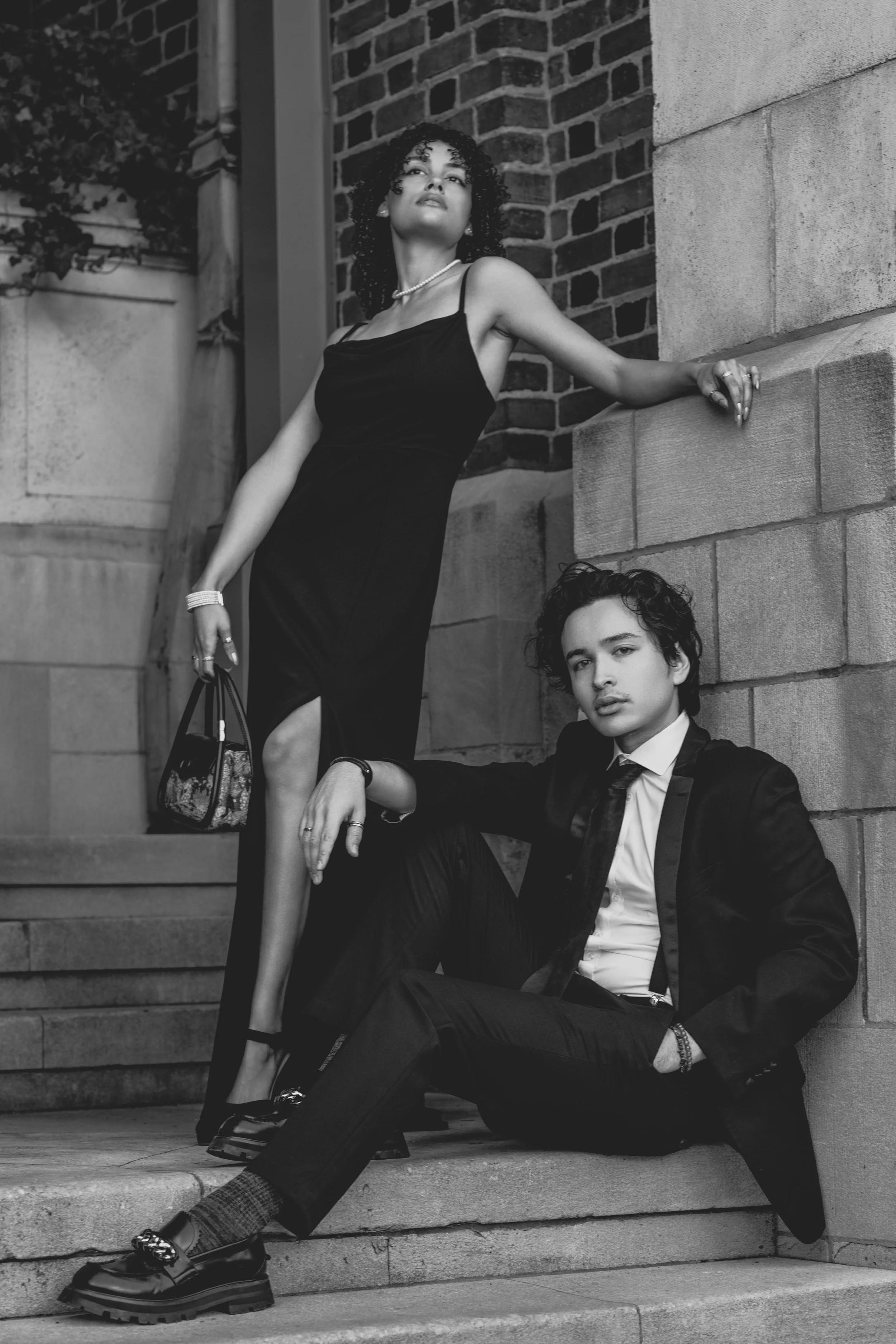 A black and white photo of a woman in a long black dress with a slit, and a man in a suit, posing on outdoor steps next to a brick wall.