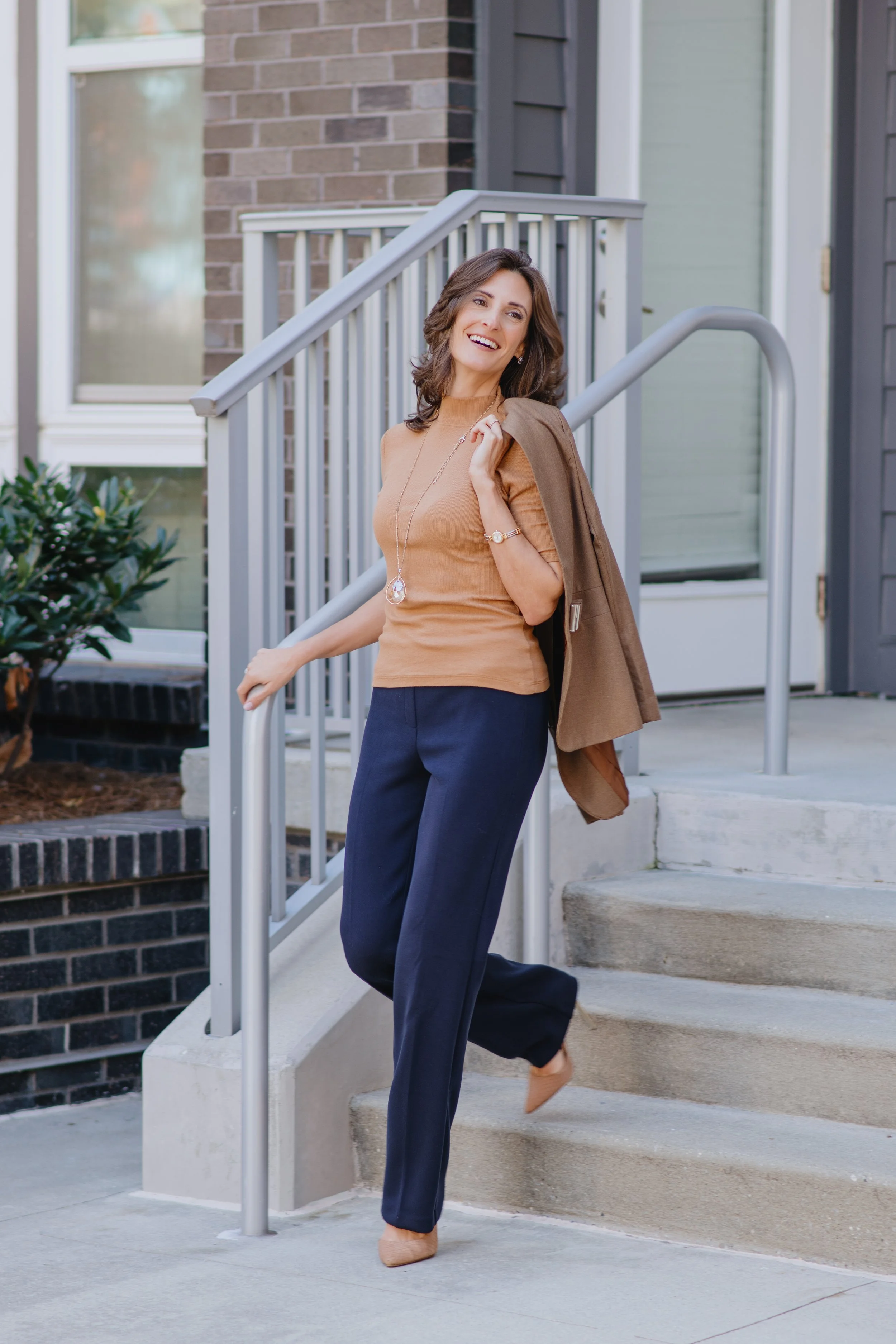 A woman smiling and holding a brown jacket over her shoulder, walking down concrete stairs outside a house with brick and siding exterior.