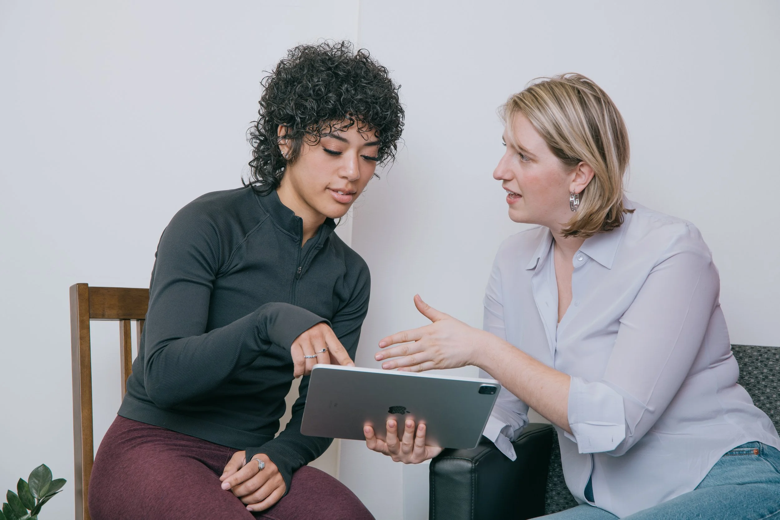 Two women sitting close together, one showing her a tablet, engaged in a conversation in a casual setting.