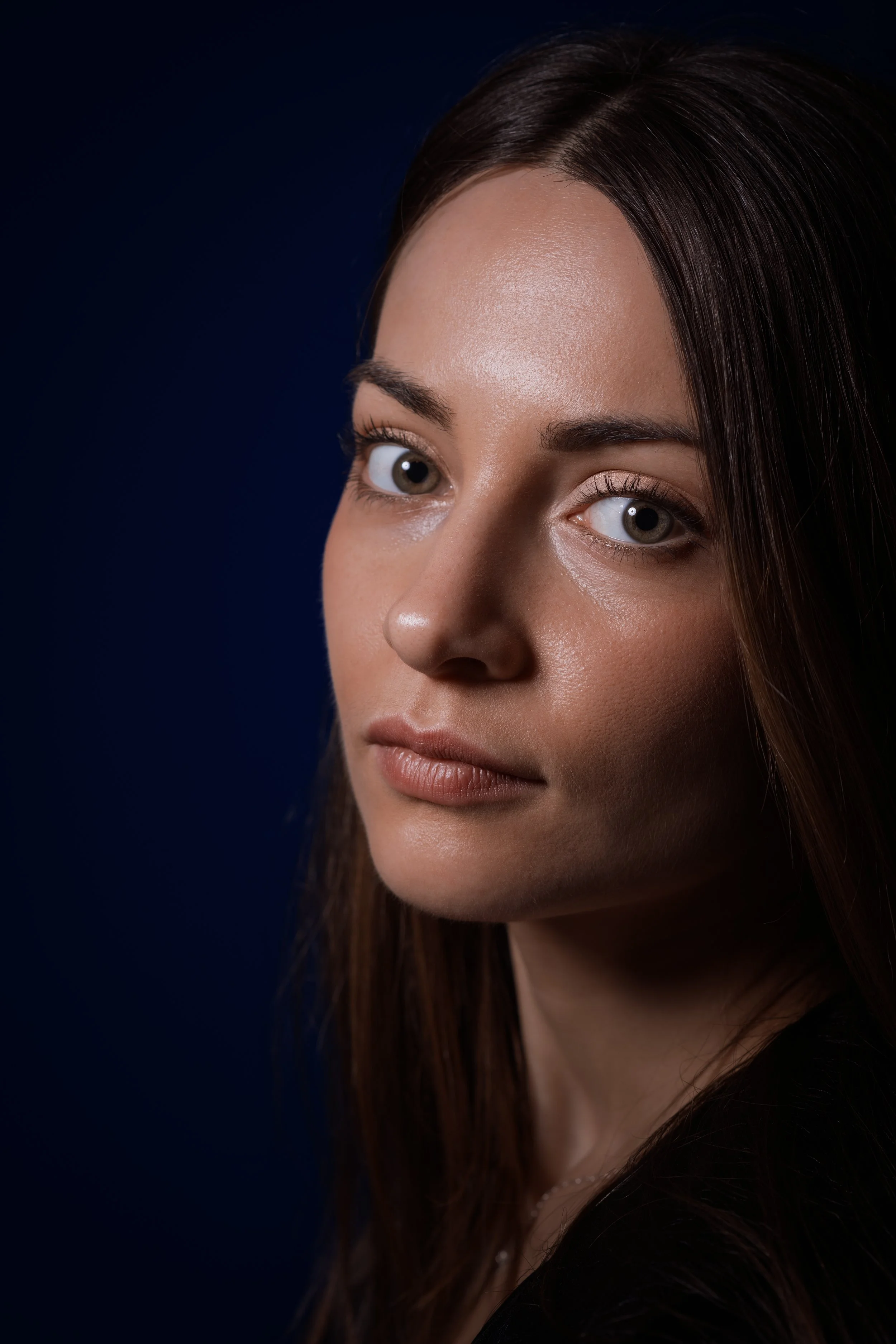 Close-up portrait of a woman with striking blue eyes and long brown hair, looking slightly to the camera against a dark background.