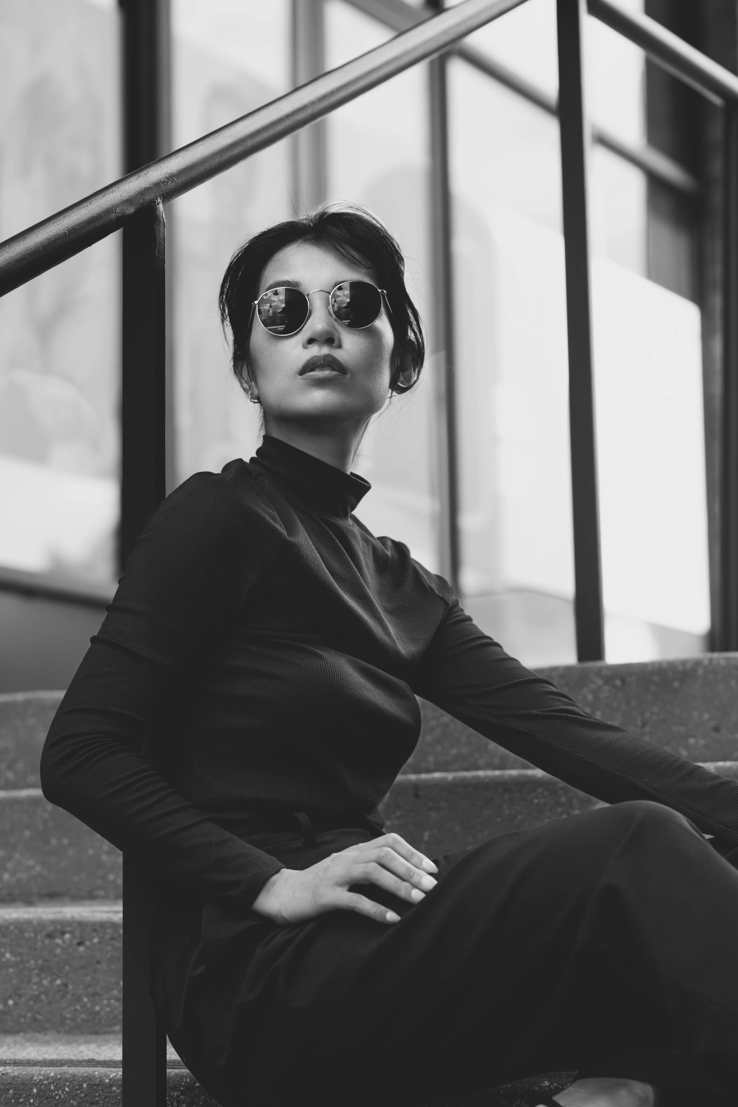 A woman wearing dark clothing and sunglasses sitting on stairs outside a building, with a reflective glass window behind her.