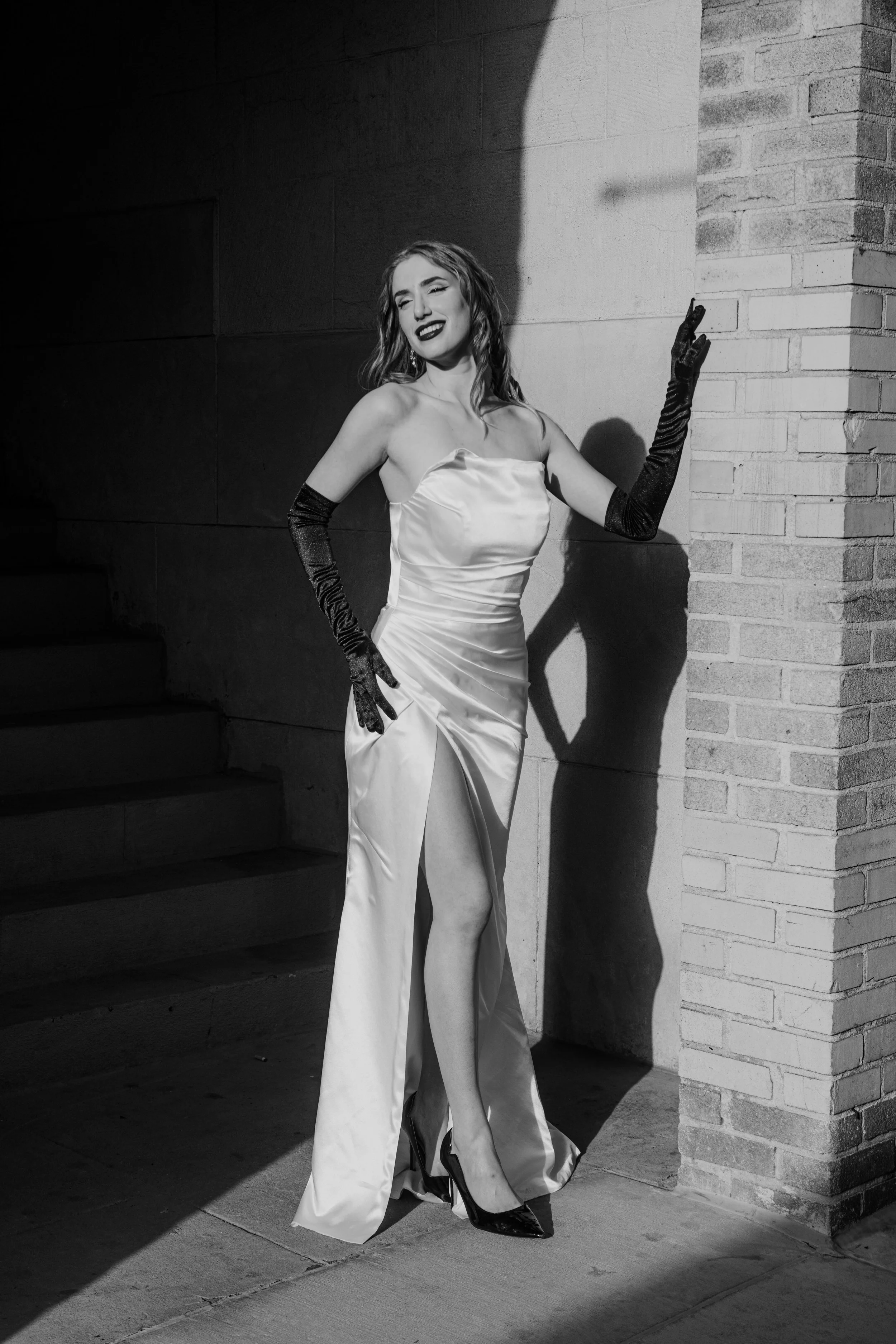 Woman in a satin gown with a high slit, wearing long gloves, smiling, standing next to a brick wall with a shadow cast on it, in black and white.