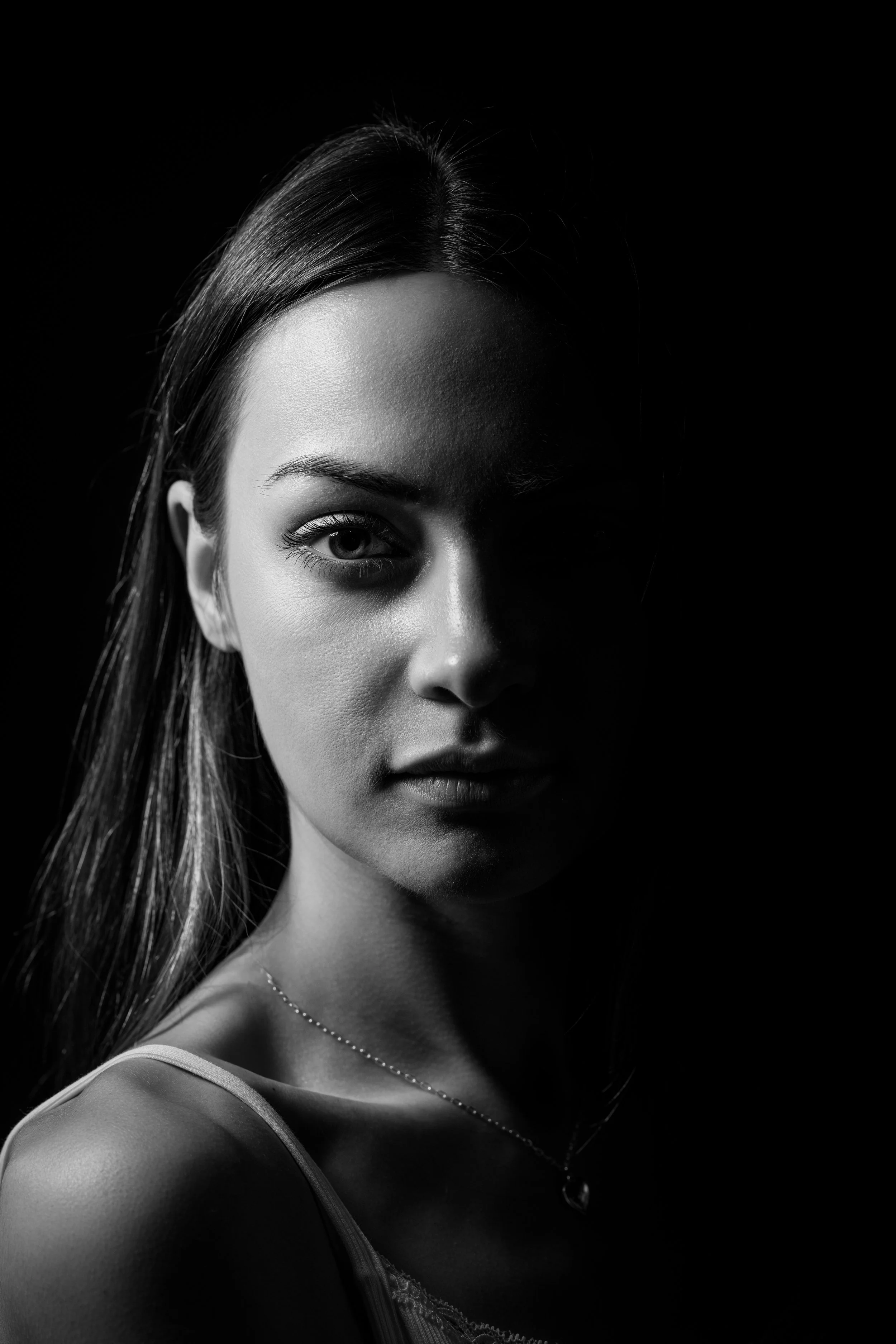 Black and white photograph of a young woman with light skin and long hair, looking directly at the camera with one side of her face highlighted and the other shadowed, wearing a single-strap top and a necklace.