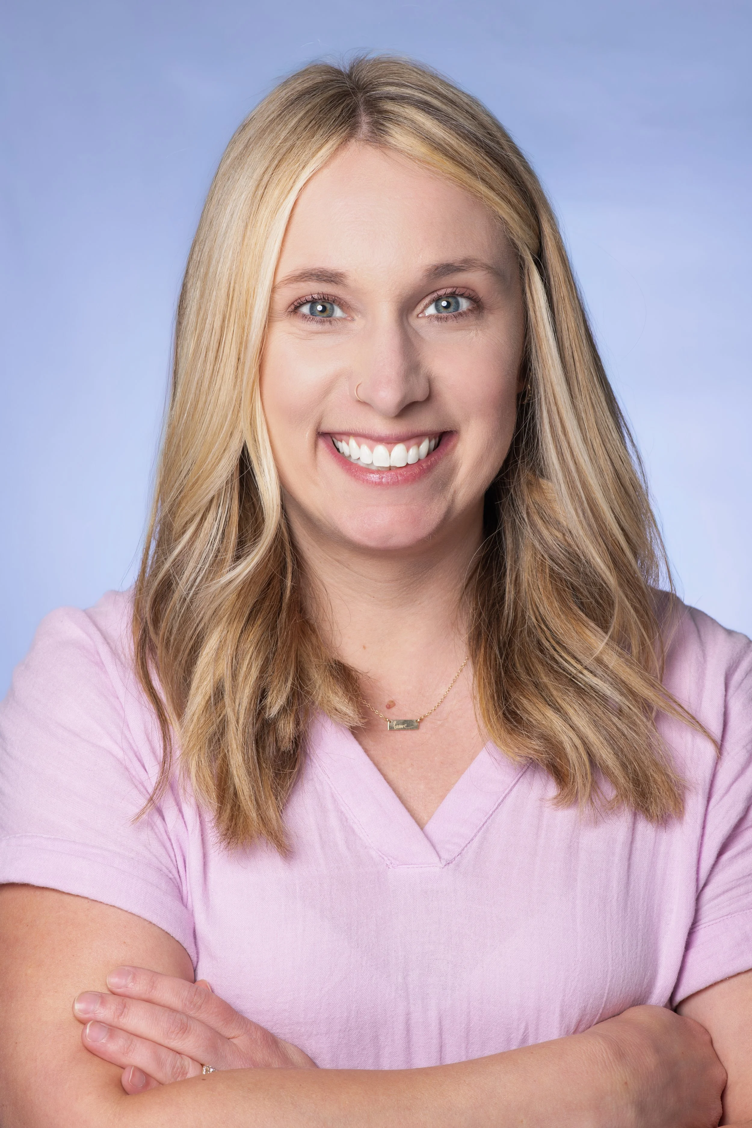 Professional headshot of a woman with a blue backdrop with her arms crossed facing camera directly. She is wearing a pink top.