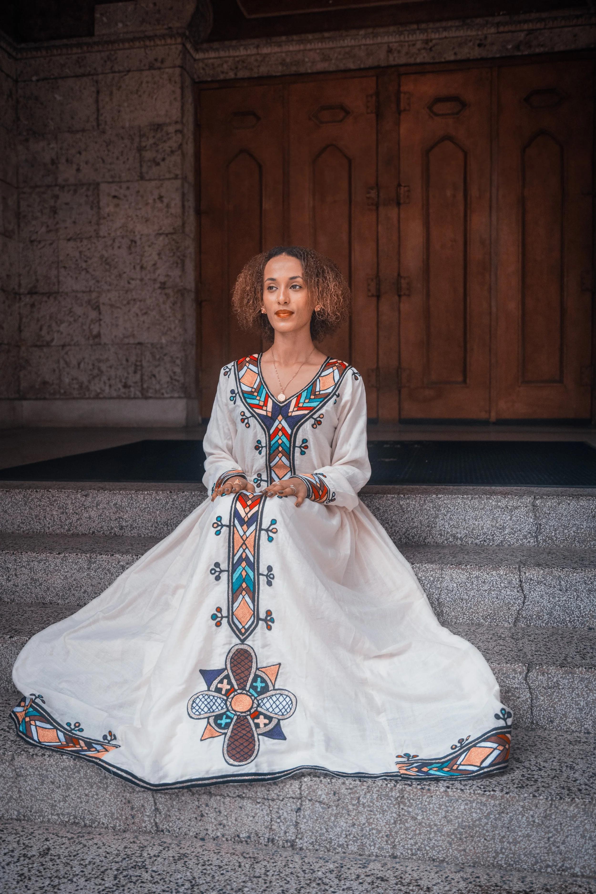 A woman sitting on steps in front of wooden doors, wearing a long white dress with embroidered colorful geometric patterns