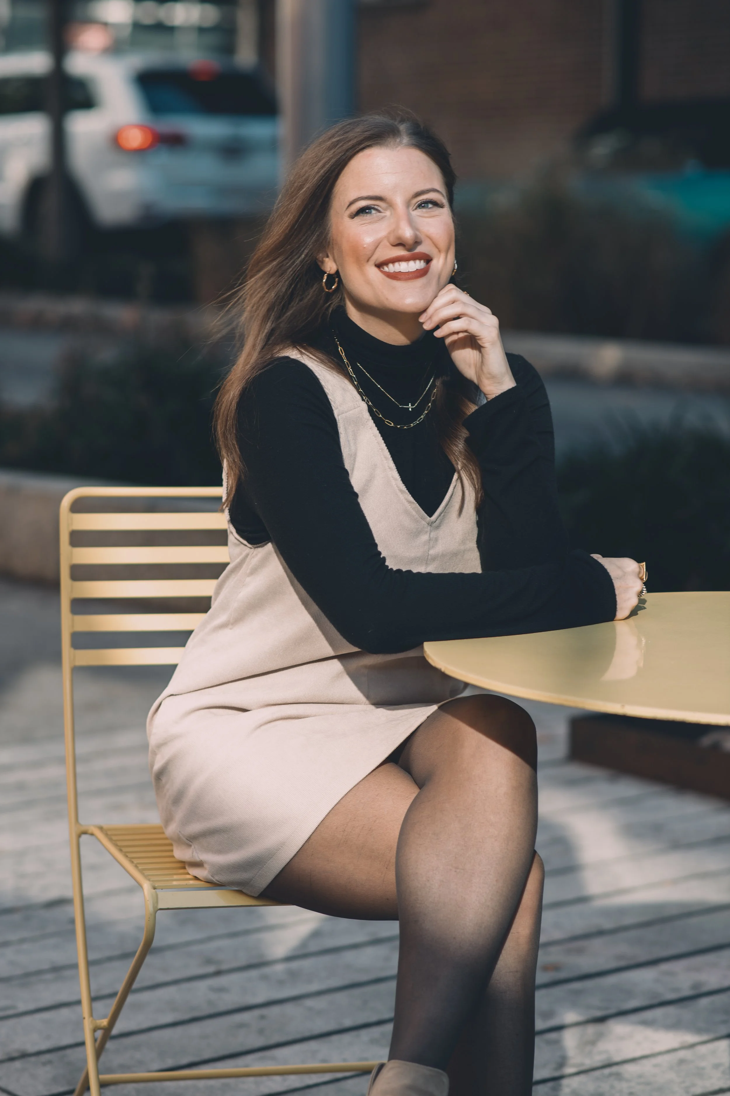 A woman with long brown hair, wearing a black turtleneck, beige dress, and black stockings, sitting at an outdoor table with a yellow chair, smiling and looking at the camera.