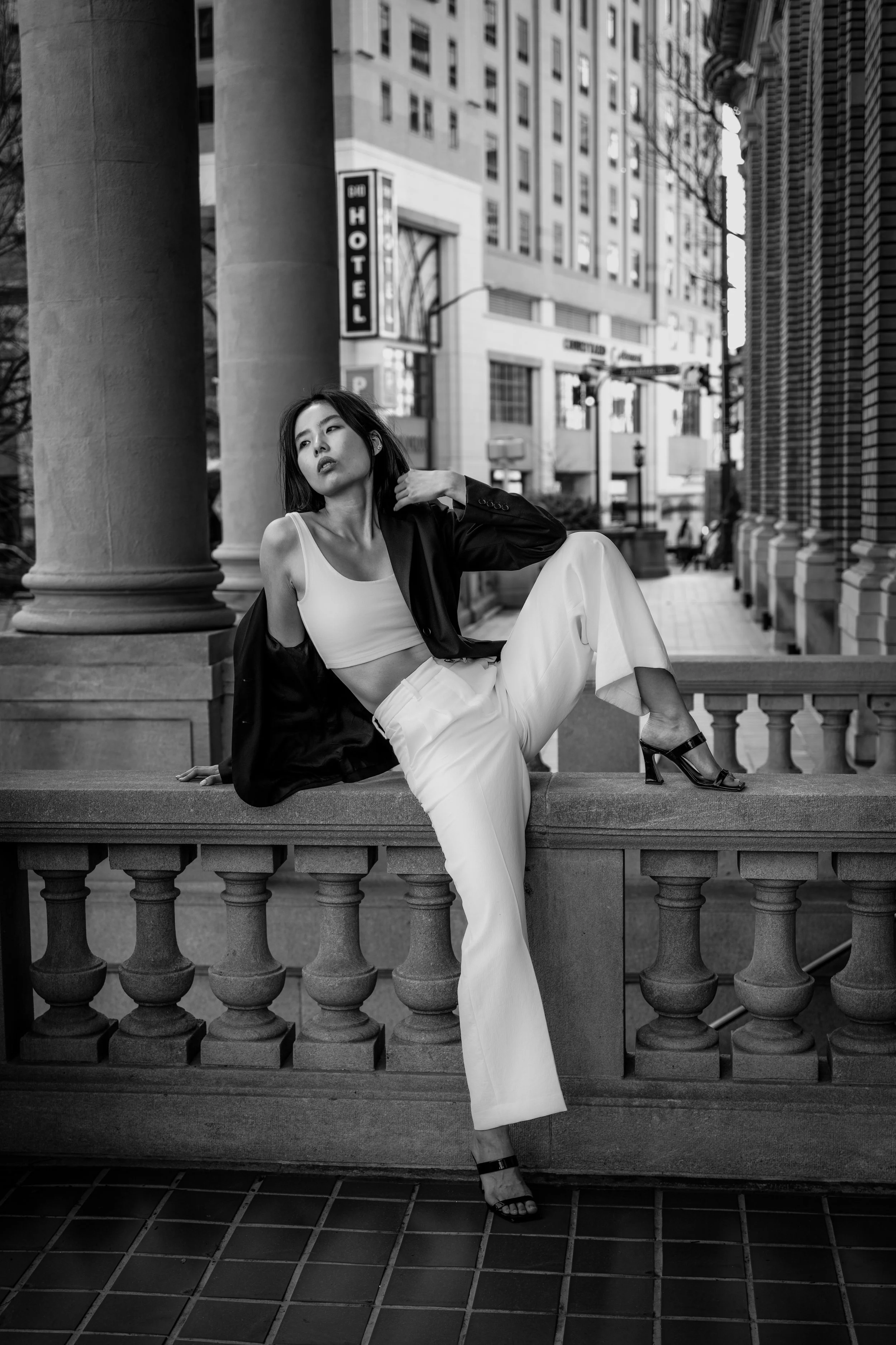 A woman in white pants and a white crop top with a black jacket draped over her shoulders sitting on a stone railing in an urban setting, with tall buildings and a hotel sign visible in the background. The image is in black and white.