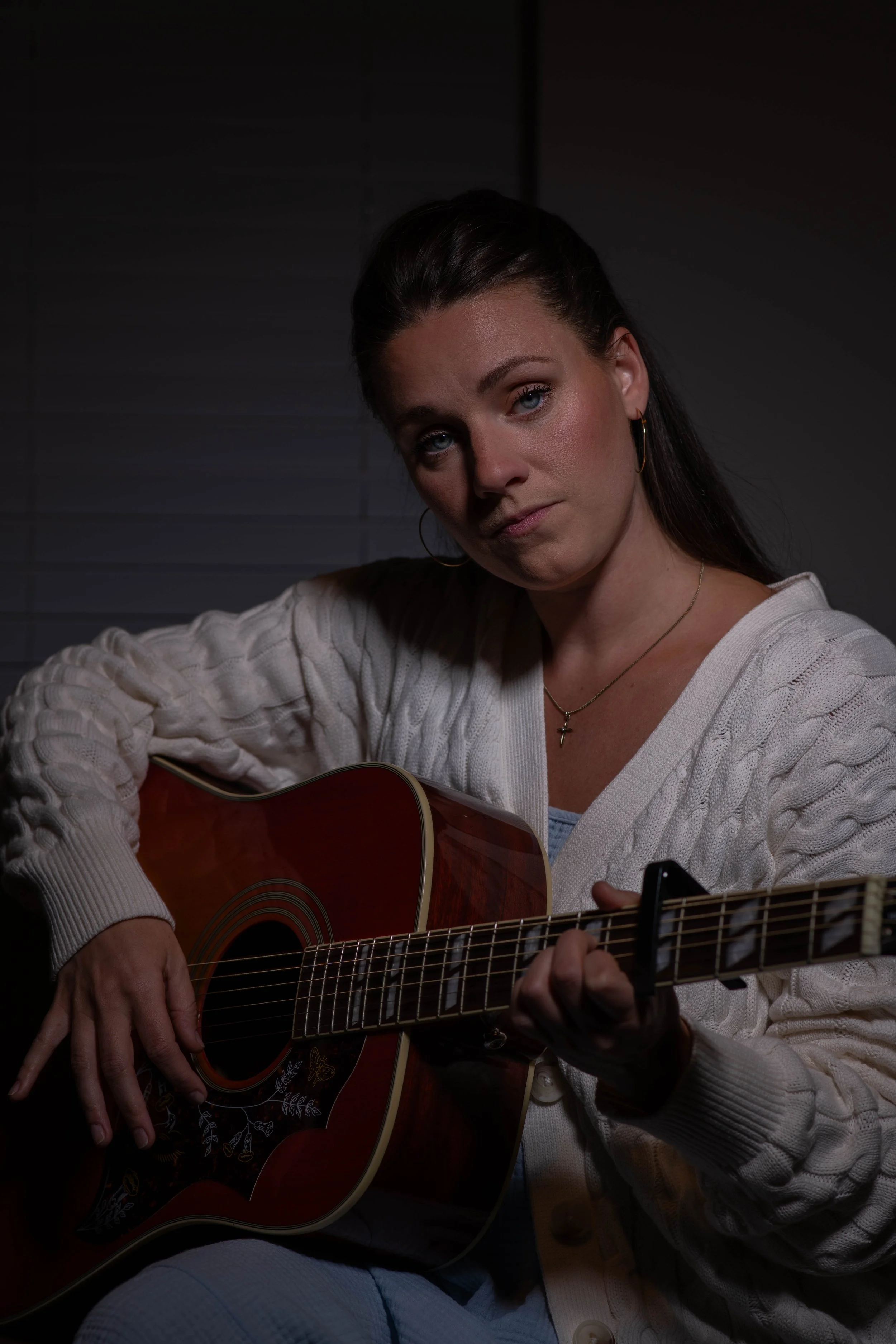 A woman with dark hair, wearing a white sweater and gold jewelry, playing an acoustic guitar in a dimly lit room.