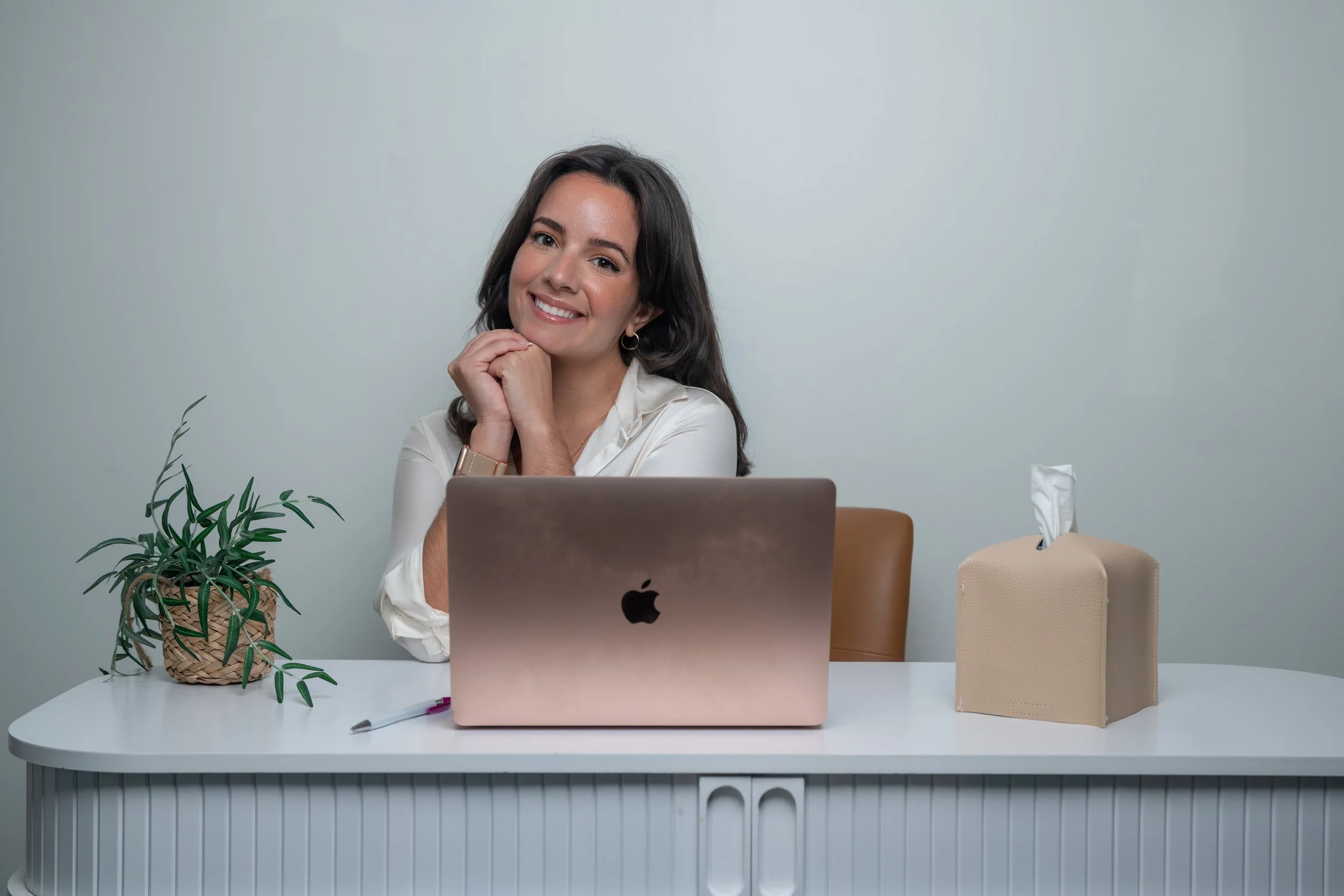 A woman sitting at a white desk with a laptop, a potted plant, a pen, and a tissue box, smiling and resting her chin on her hands.