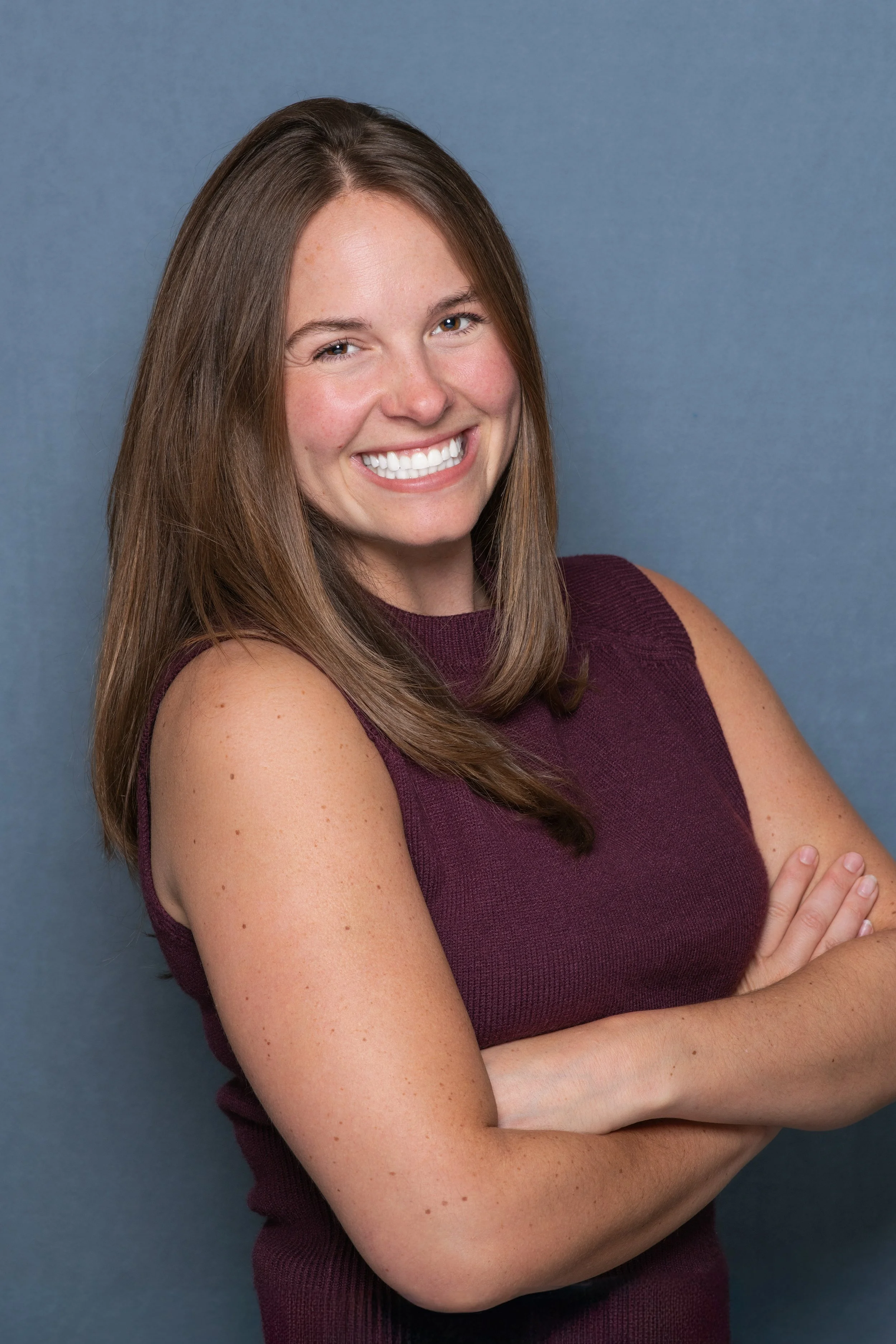 Professional headshot of a woman with a blue backdrop with her arms crossed facing camera right.