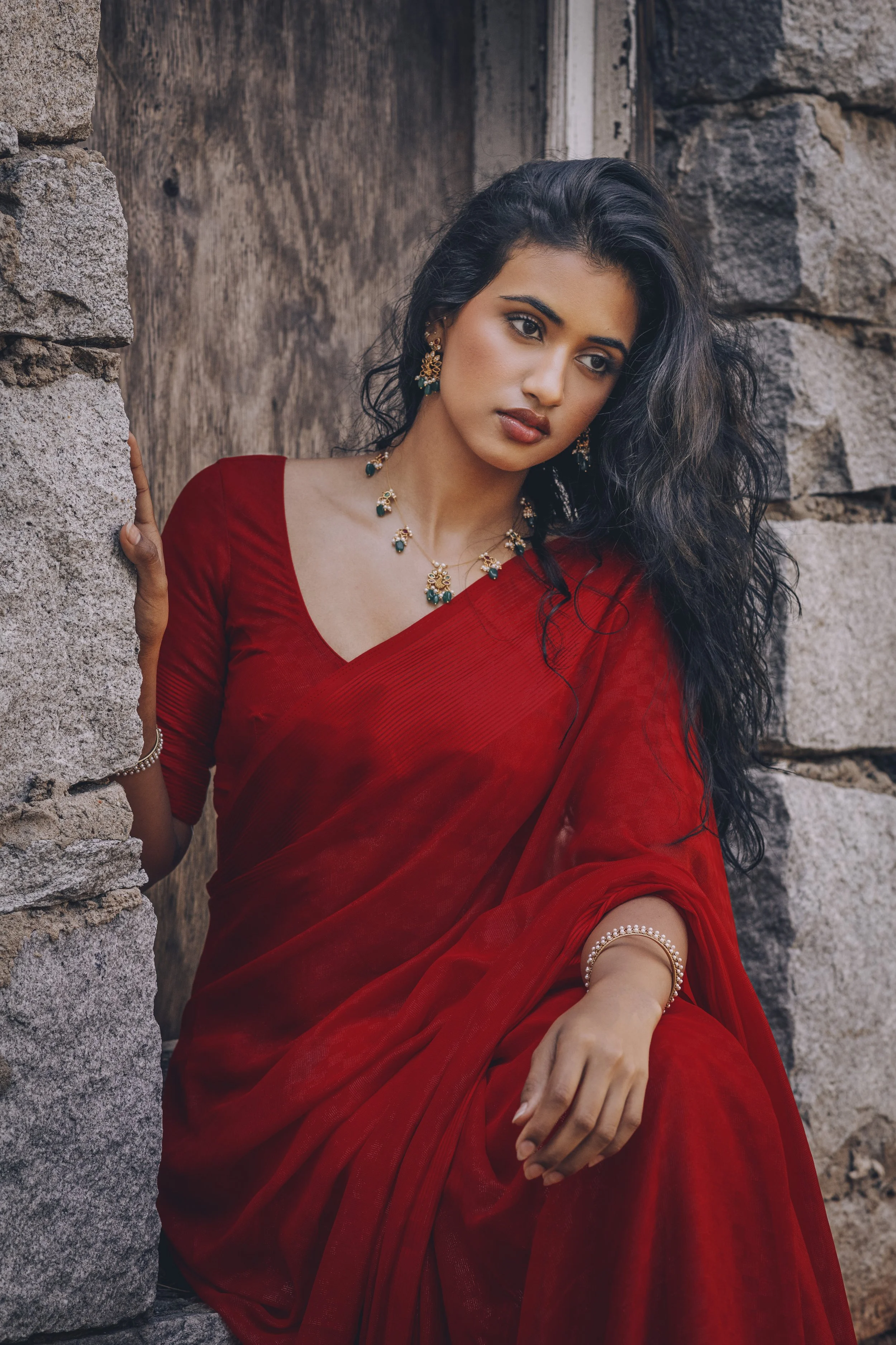 A woman in a red sari with dark hair, wearing gold and gemstone jewelry, sitting against a stone wall.