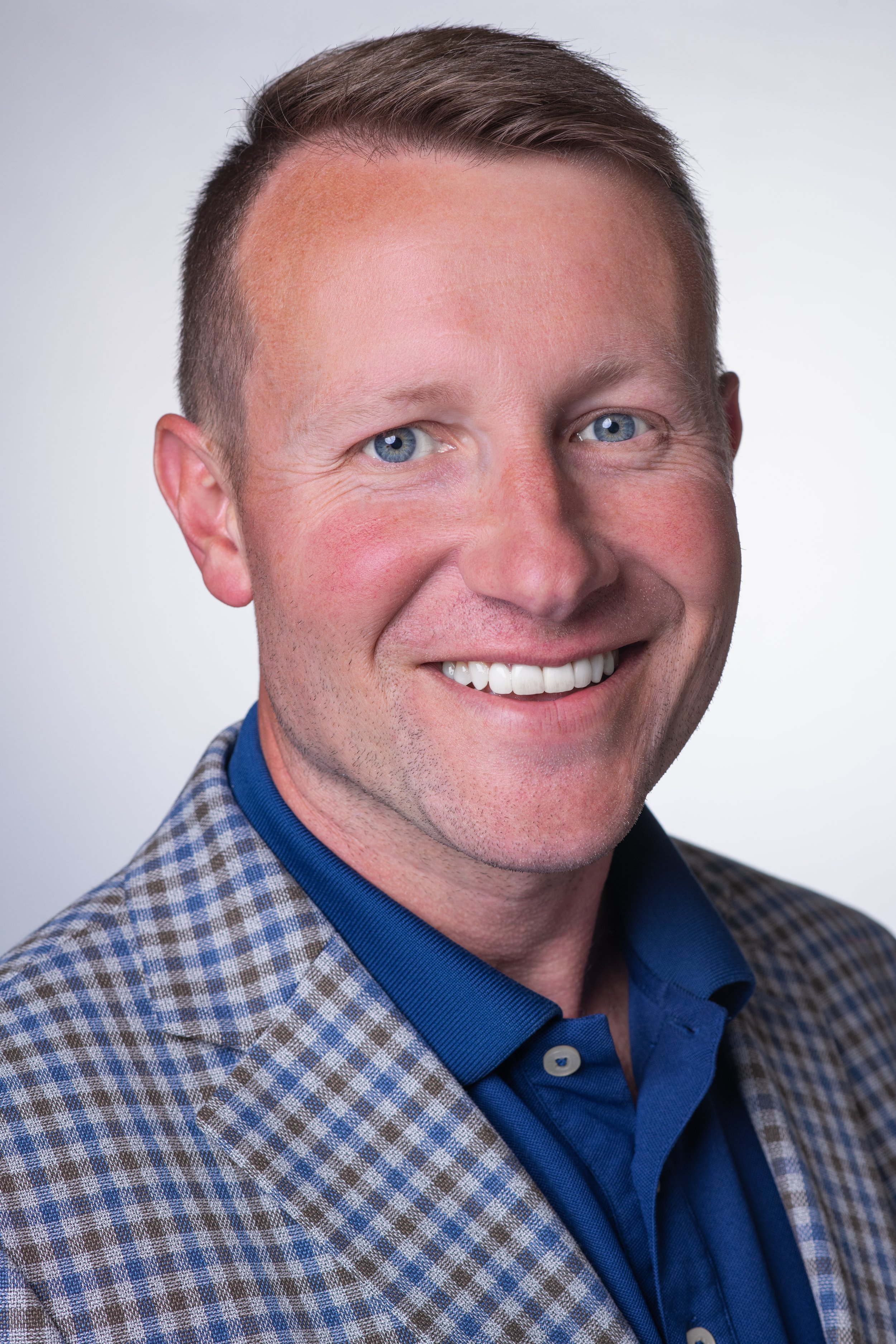Professional headshot of a man with a white backdrop turned towards camera right but looking towards the camera. He is wearing a blue check patterned suit.