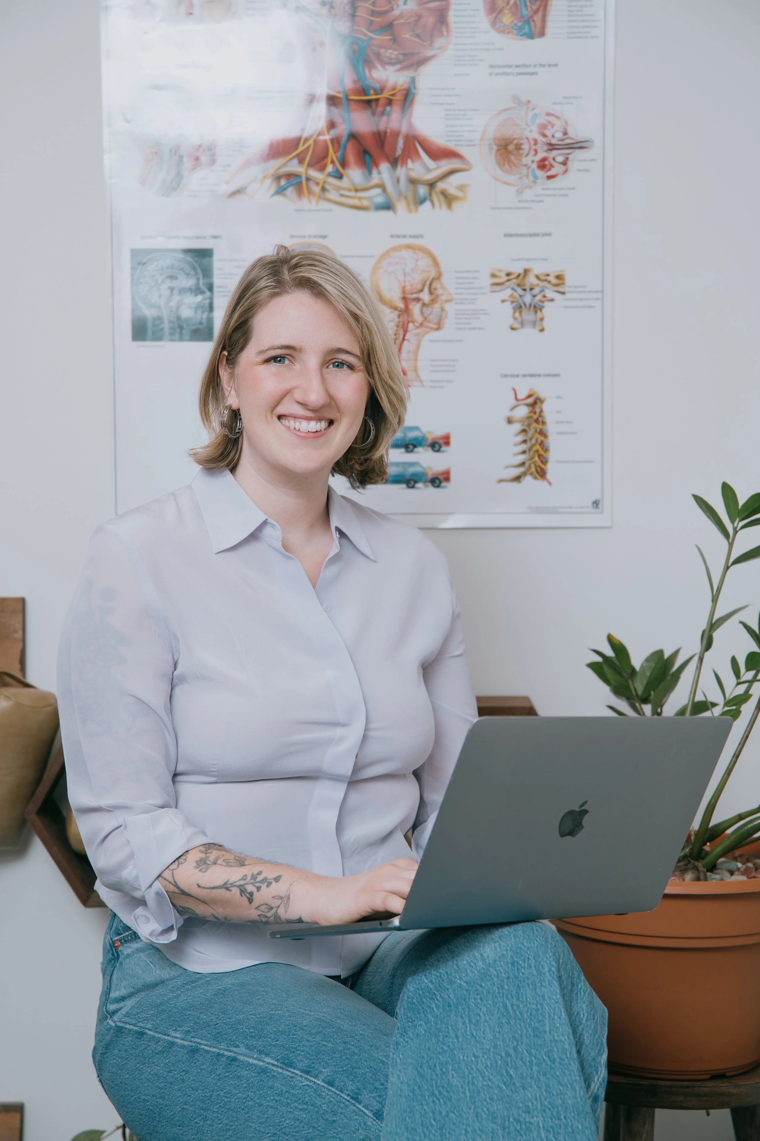 A woman sitting on a stool with a laptop, smiling, with a poster of the human nervous system behind her and a potted plant nearby.