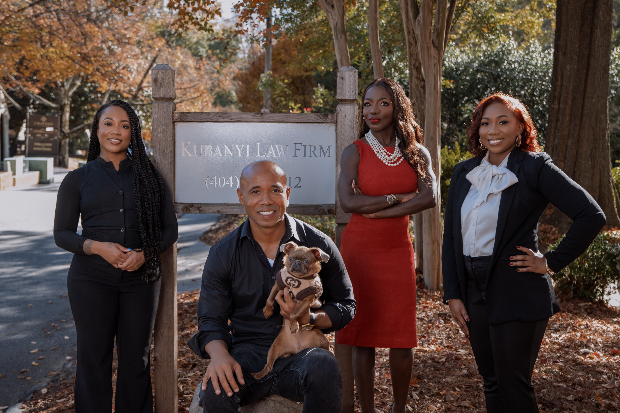 Group of four diverse adults and one dog standing outdoors in front of a sign for Kubanyi Law Firm, with trees displaying autumn foliage in the background.