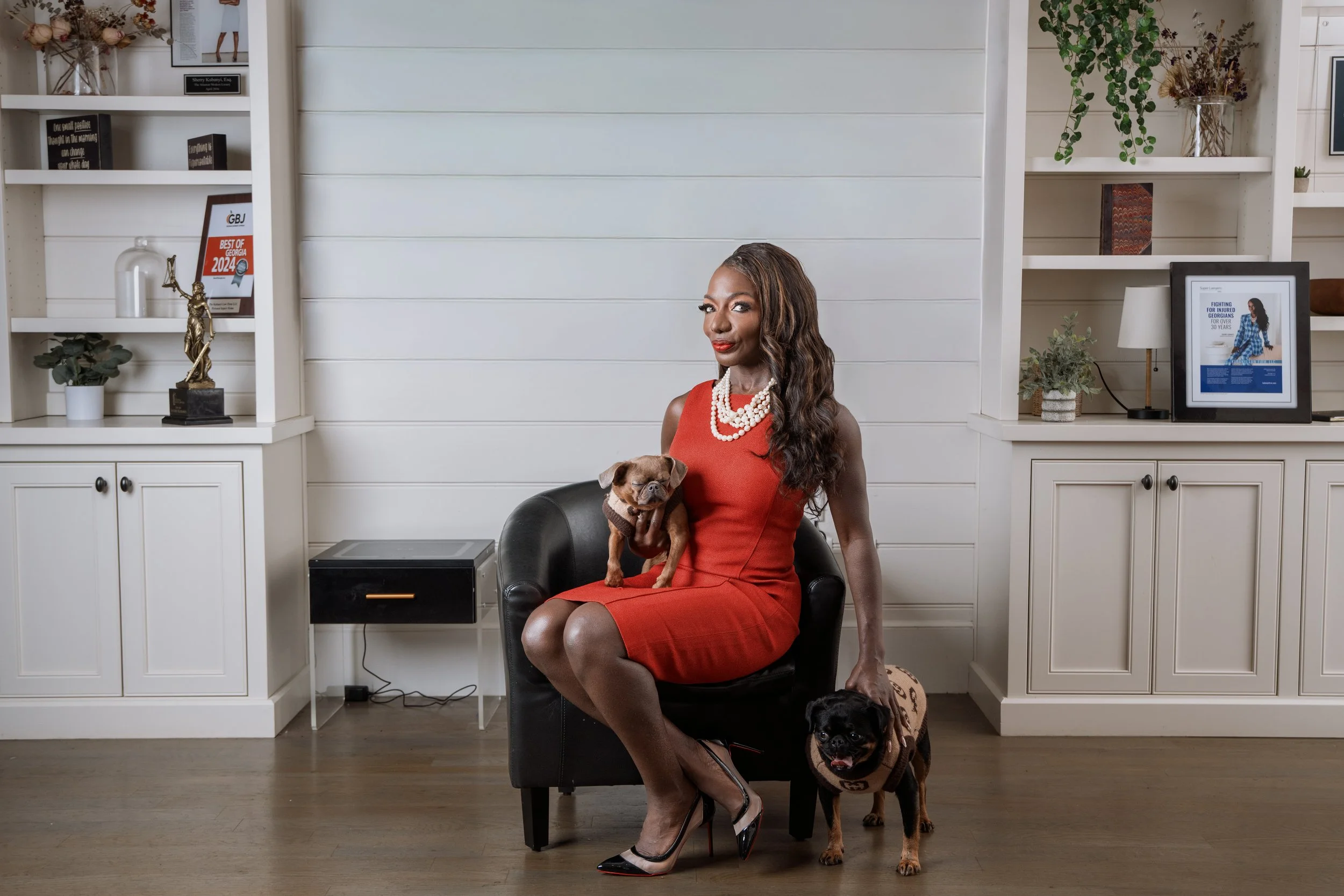 A woman in a red dress with pearl necklaces sitting on a black armchair in a well-decorated room, holding a small dog on her lap and a larger dog standing next to her.
