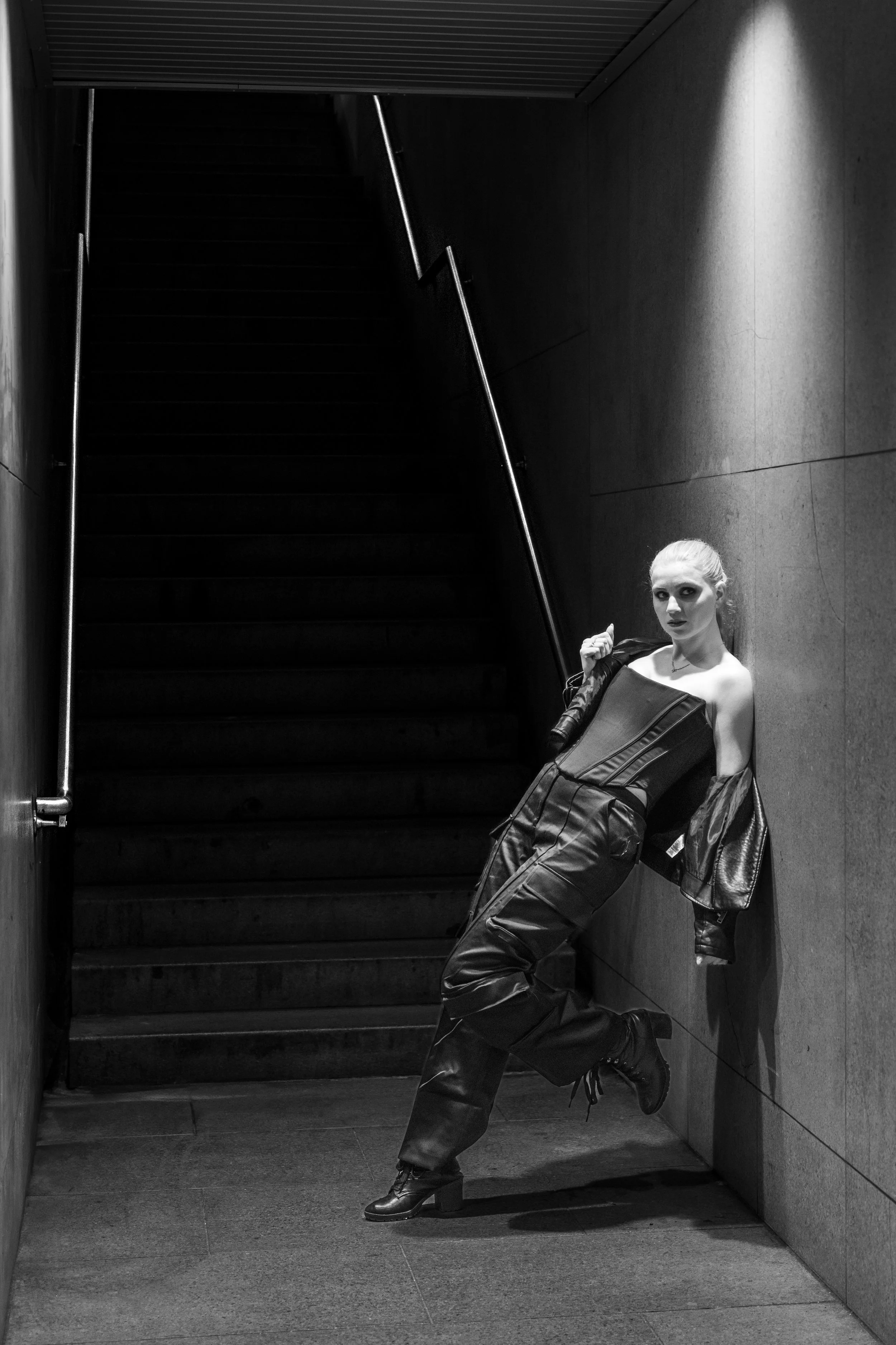 Black and white photo of a woman leaning against a wall in a stairwell, wearing leather pants, heels, and a corset, with a leather jacket draped over her shoulders.