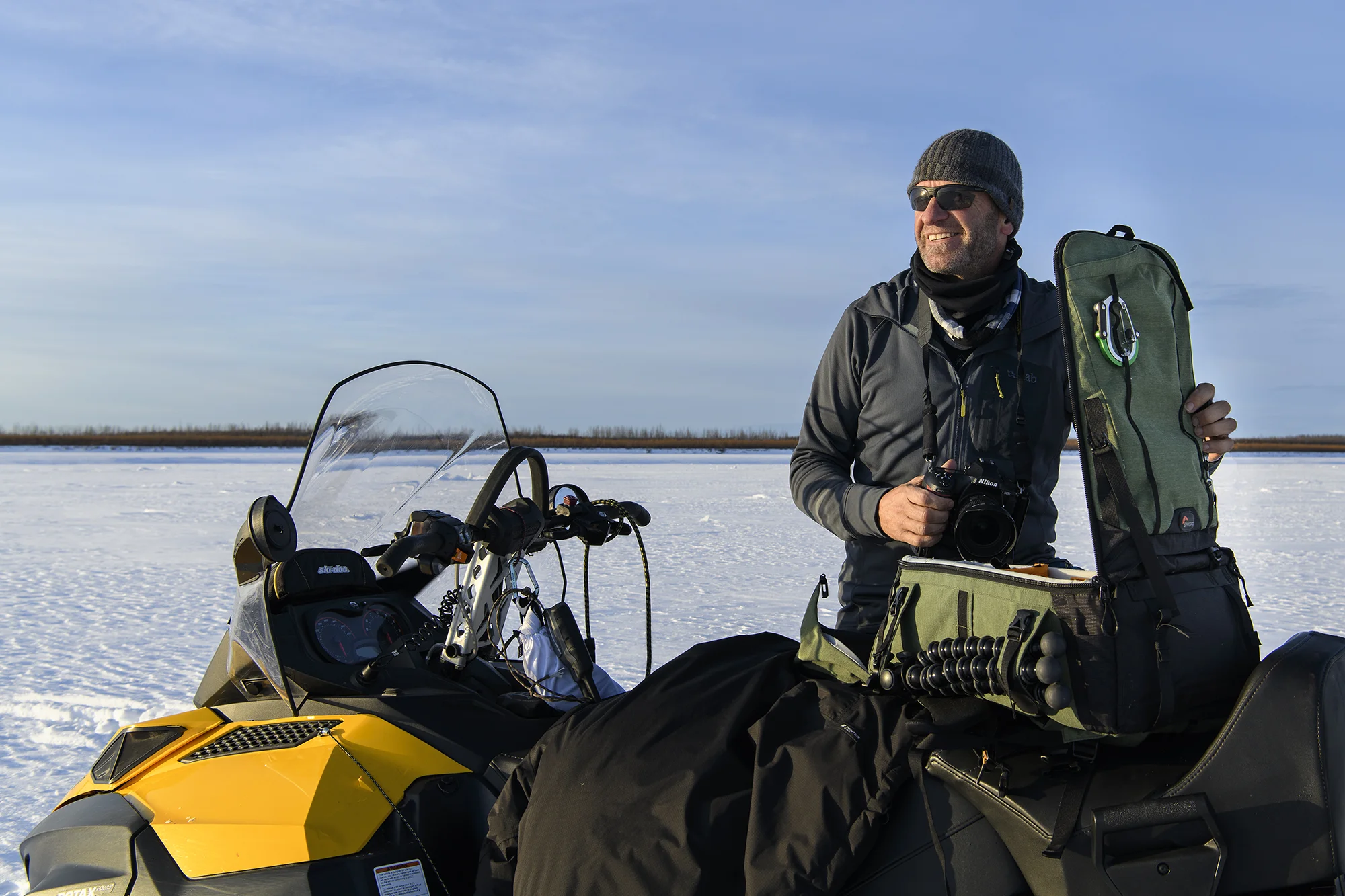 A man dressed in winter gear, including a knit hat and sunglasses, stands outdoors on snow with a yellow snowmobile and photography equipment, holding a camera bag and smiling in a wintry landscape under a clear blue sky.