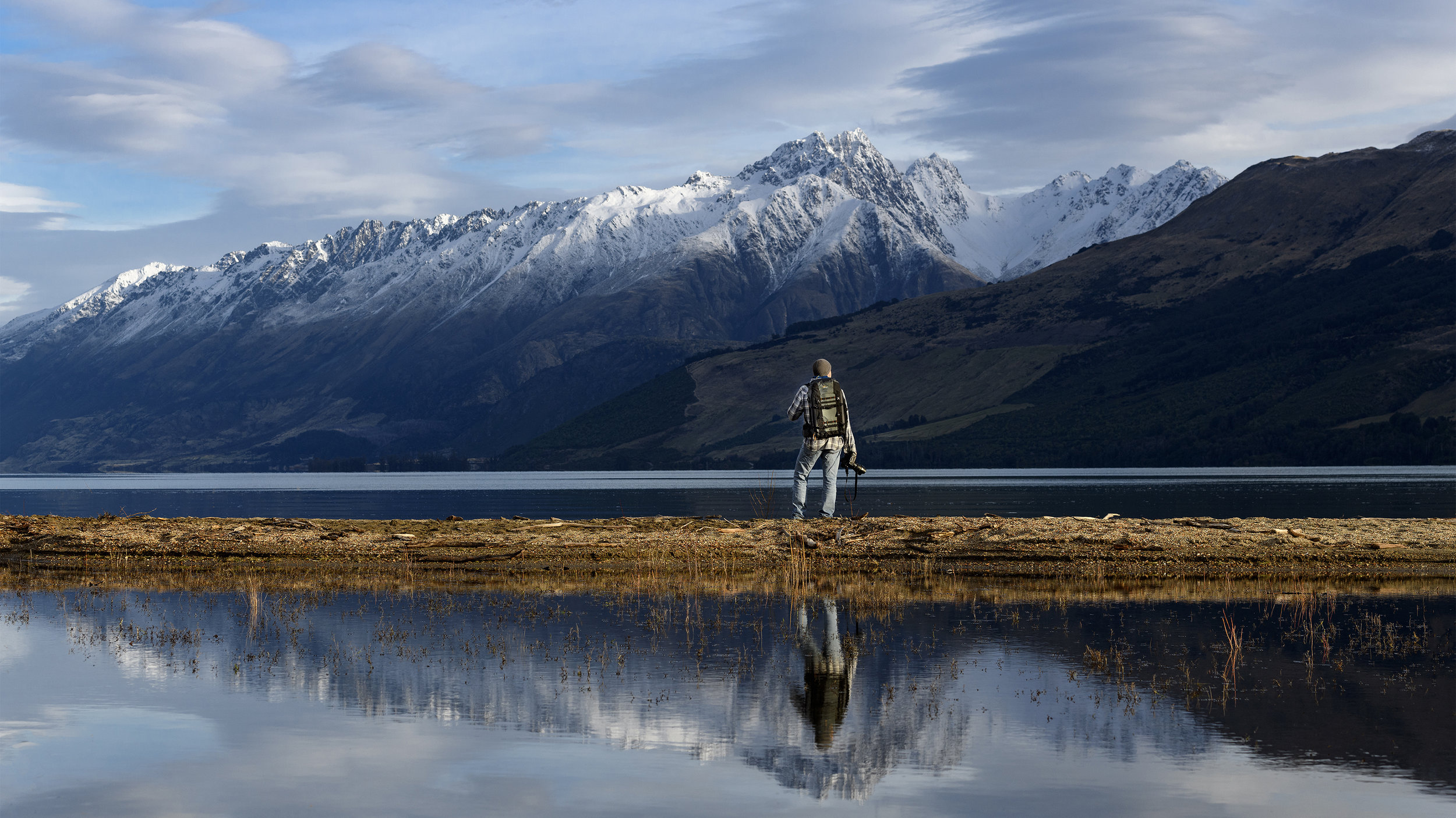 Chris McLennan standing on Lake Wakatipu in Glenorchy with a Lowepro a backpack on the shore of a lake wakatipu, looking at snow-capped mountains.