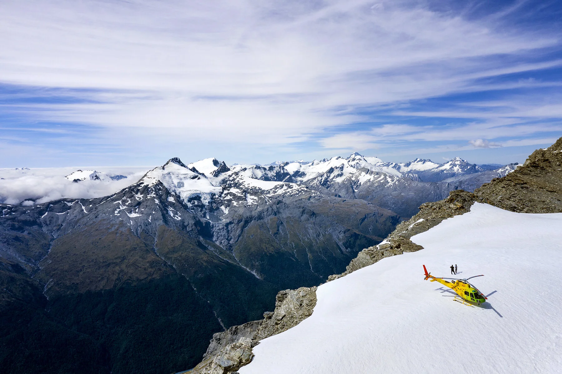 A helicopter parked up on a glacier with two people standing by looking at the landscape of the new zeland southern alps.