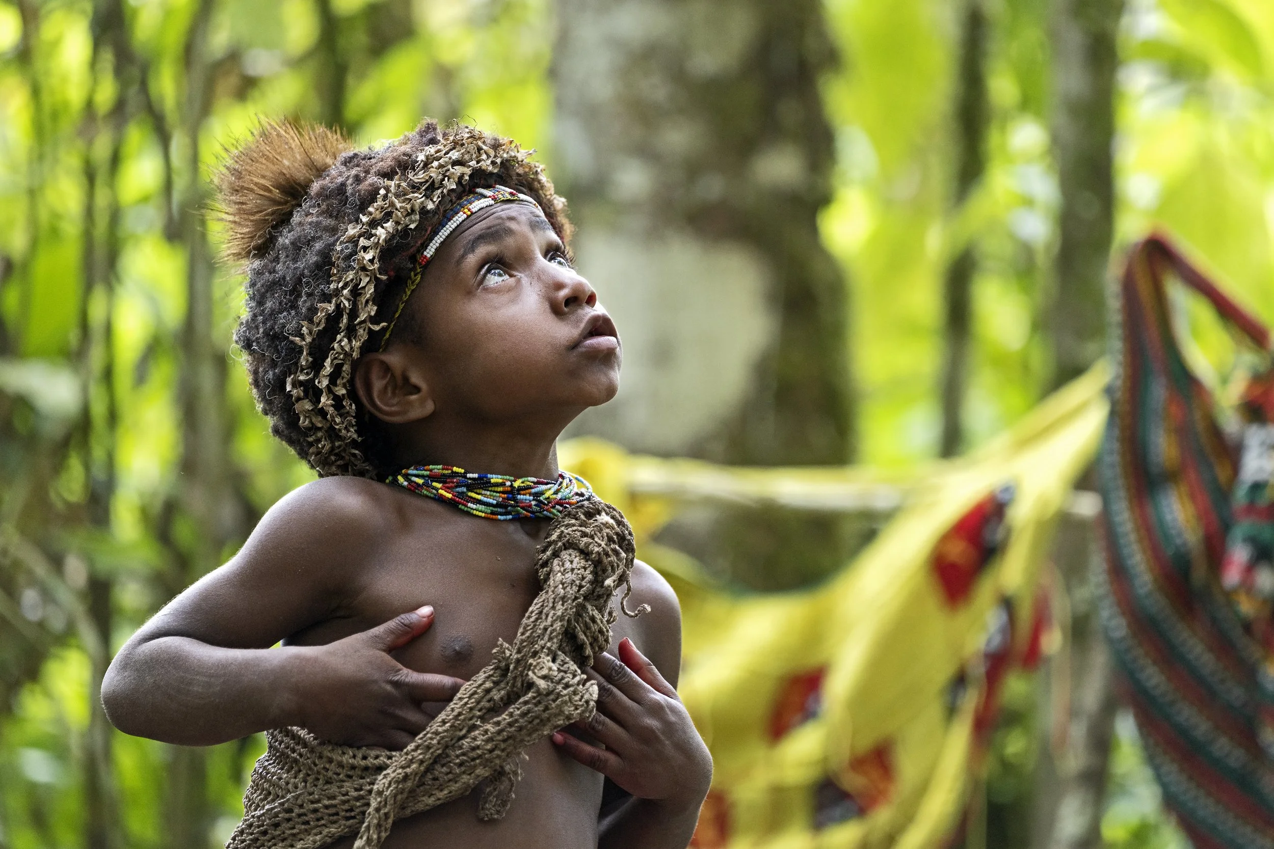 A young person in Paupa New Guinea with traditional jewelry and headwear looking upward while holding a rope with one hand and placing the other hand on their chest, in a lush green forest.