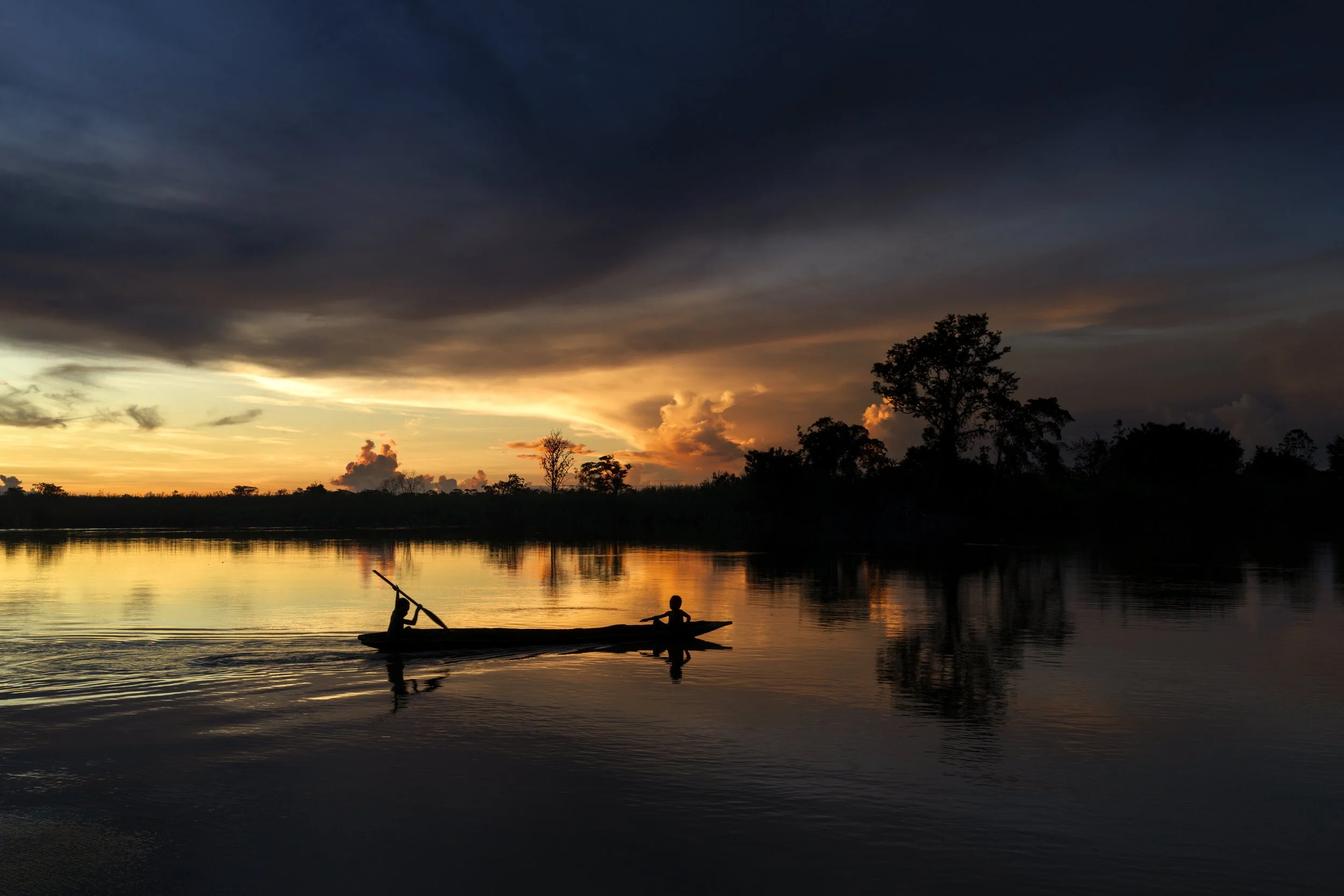 Silhouetted figures in a boat on a calm river at sunset with a darkening sky and trees in the background.