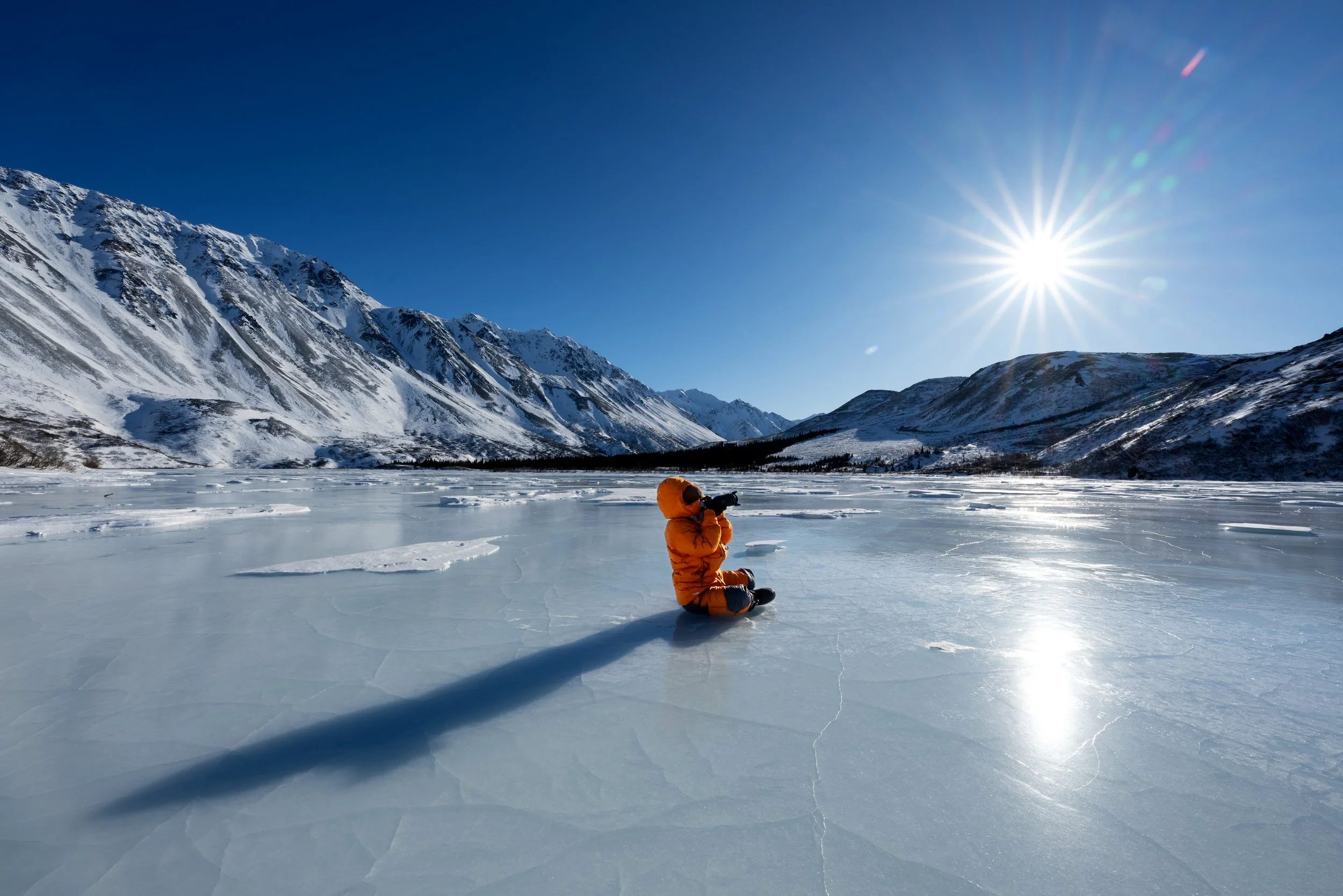 A person in an orange winter coat kneeling on snow-covered ice, holding a camera, with snow-capped mountains in the background and the sun shining brightly in a clear blue sky.