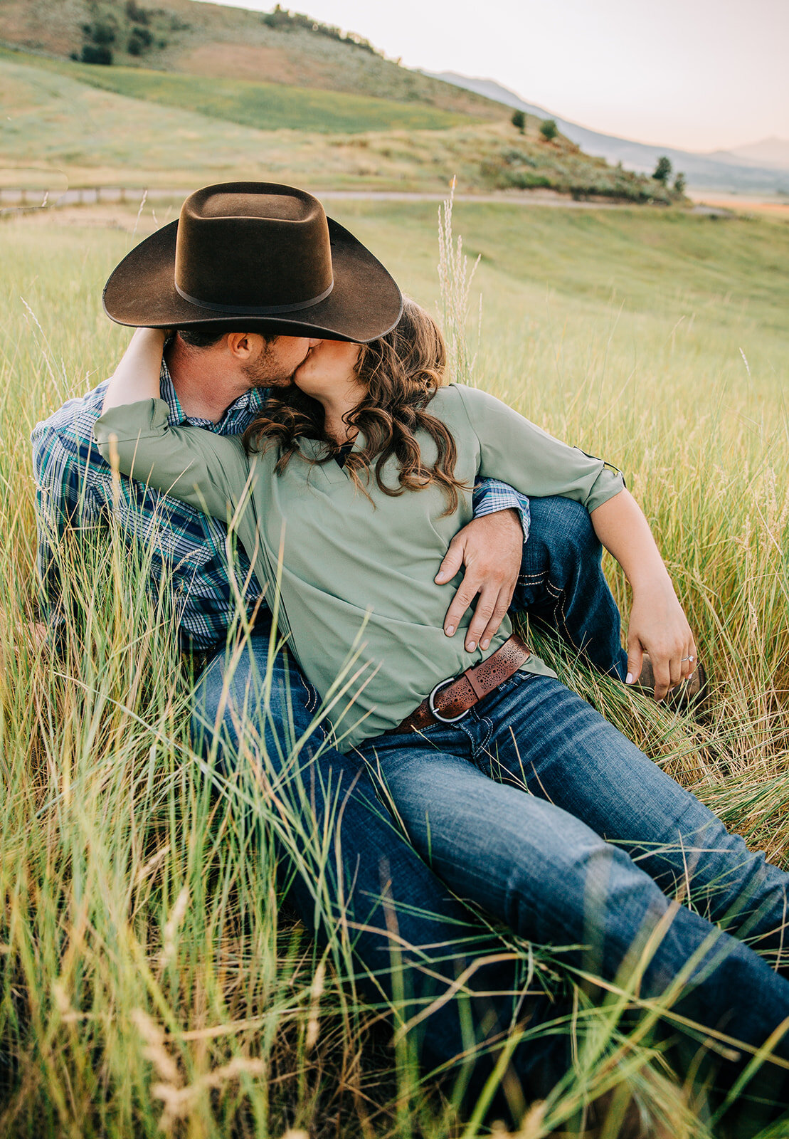  couple sitting in grass field green background engagement session poses intimate engagement session kissing photo inspo happy couple engagement outfit inspo blue jeans relaxed photoshoot country couple small town love cowboy dancing partners cache v