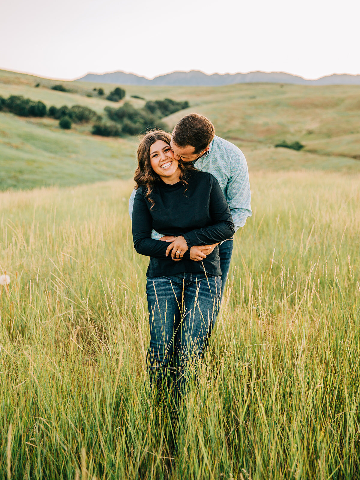  country love small town couple summer infused engagement session green hills and grass fields hugging poses romantic posing hands around waist kissing pictures big smiles men and women hairstyles engagement outfit inspo cache valley photographer par