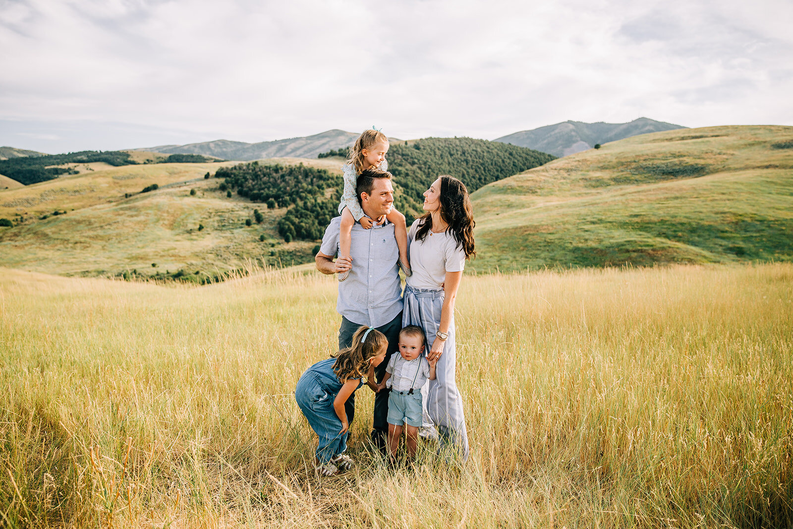  small family in a yellow field with green rolling hills behind paradise utah hyrum utah professional family photographer cache valley shades of blue outfits matching color scheme for summer family photos dad with daughter on his shoulders holding ha