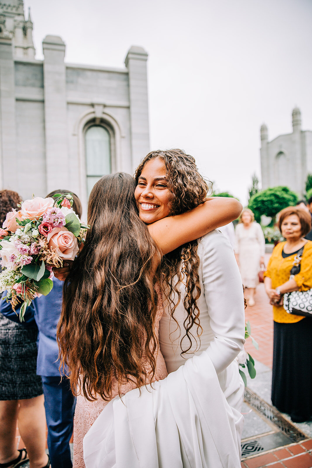  bride hugging her sister after exiting the salt lake lds temple on temple square friends and family photos after temple sealing lds bride curly hair bride smile style inspiration for young mormon brides modest bridal gown with long sleeves fall wedd