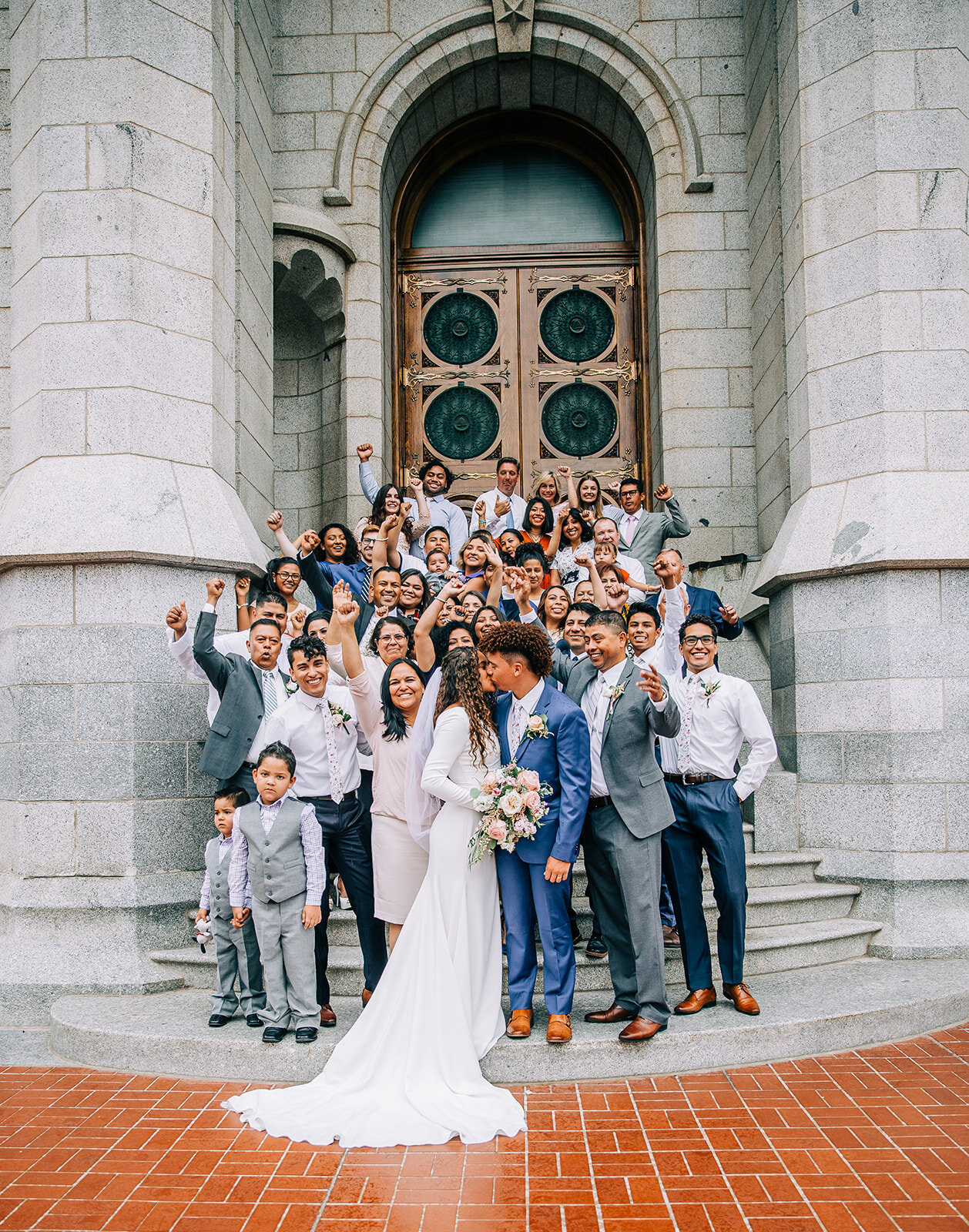  bride and groom kissing in front of family cheering on temple steps salt lake city temple square utah mormon bride lds couple long veil modest wedding gown with long sleeves winter wedding inspo neutral color scheme blue suit for the groom husband a