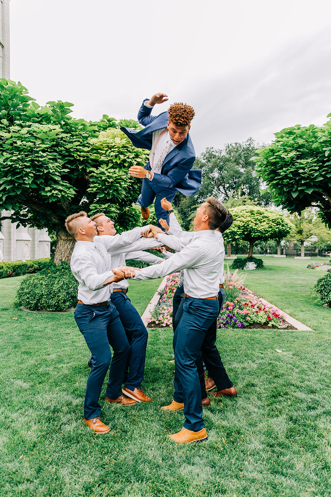  pose idea groomsmen throwing groom up in the air wedding day pictures of groomsmen tossing groom blue suit navy suit pants wedding party attire ideas inspo for wedding style blue pants with white shirts and floral ties young couple lds bride and gro