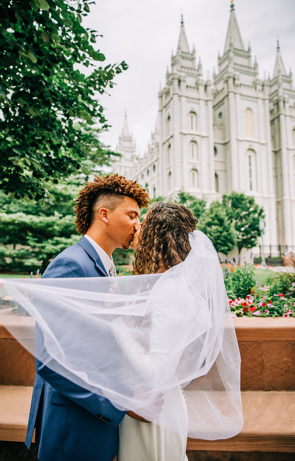  veil shot bride and groom kissing behind the veil bridals at temple square outside the salt lake city temple winter wedding bridal ensemble inspo long veil curly hair bride and groom hair ideas style inspiration for young couple wedding blue suit te