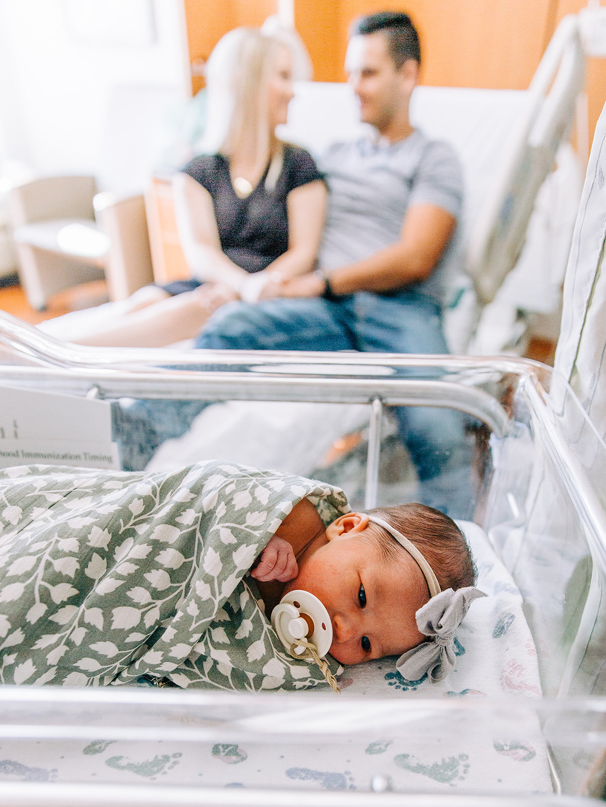  close-up newborn photo shot of baby in a grey swaddle in hospital cradle with mom and dad in the background cute pose idea for newborn photography professional birth story photographer bella alder photography fresh 48 cache valley utah affordable ne